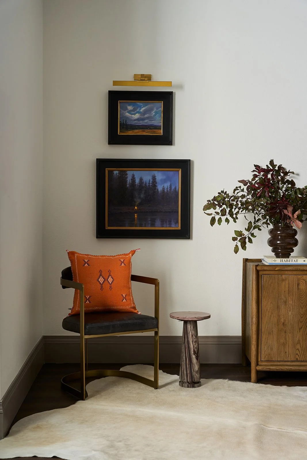 A cozy corner with a black and gold chair that has an orange patterned pillow, a small rustic wooden stool, a wooden cabinet with books and a large brown vase with greenery, two landscape paintings on the wall, and a white rug on the dark wood floor.