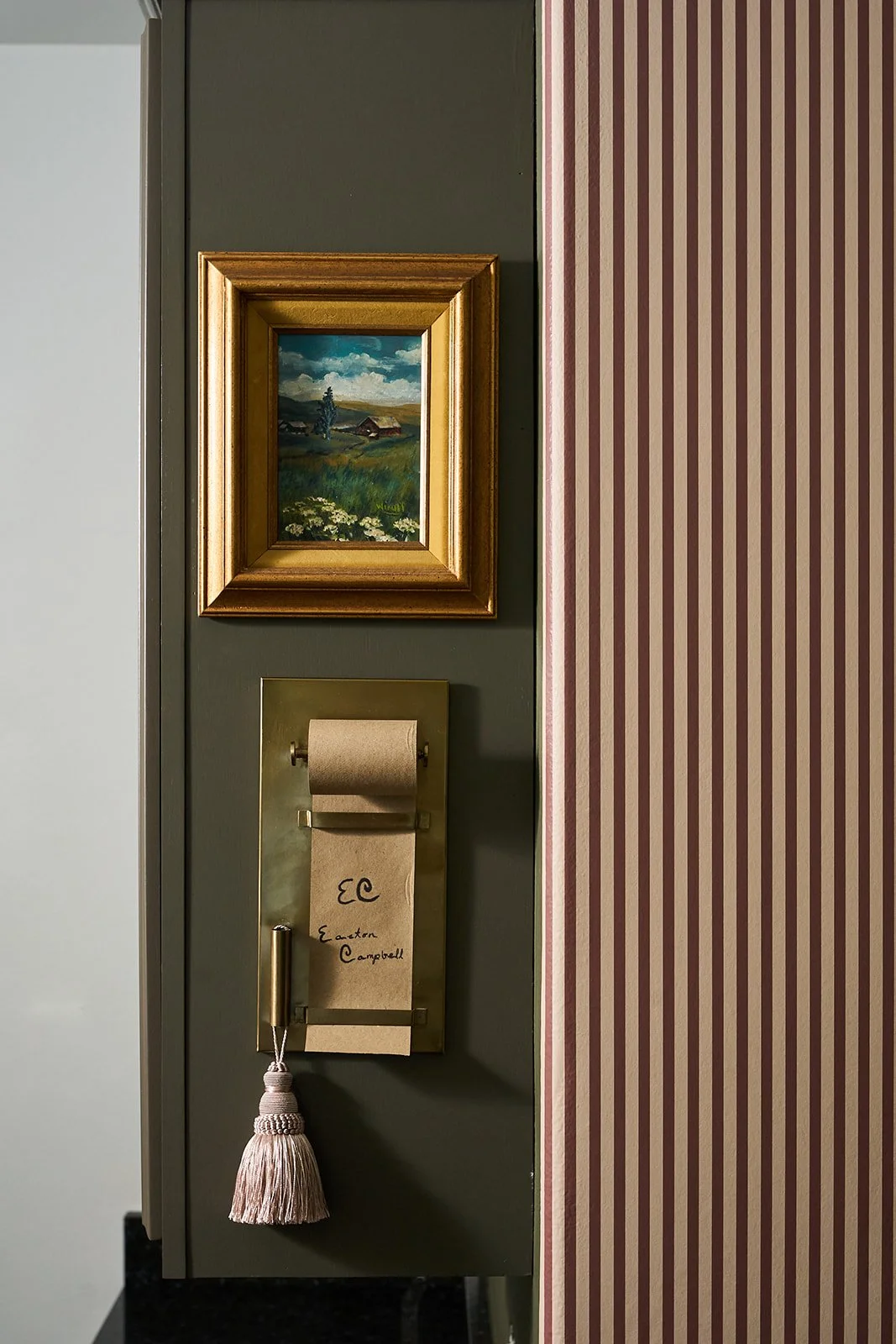 A wall with a framed landscape painting of a rural scene, a gold mailbox with a handwritten note, and a pink tassel hanging from it. The wall has dark and striped wallpaper sections.