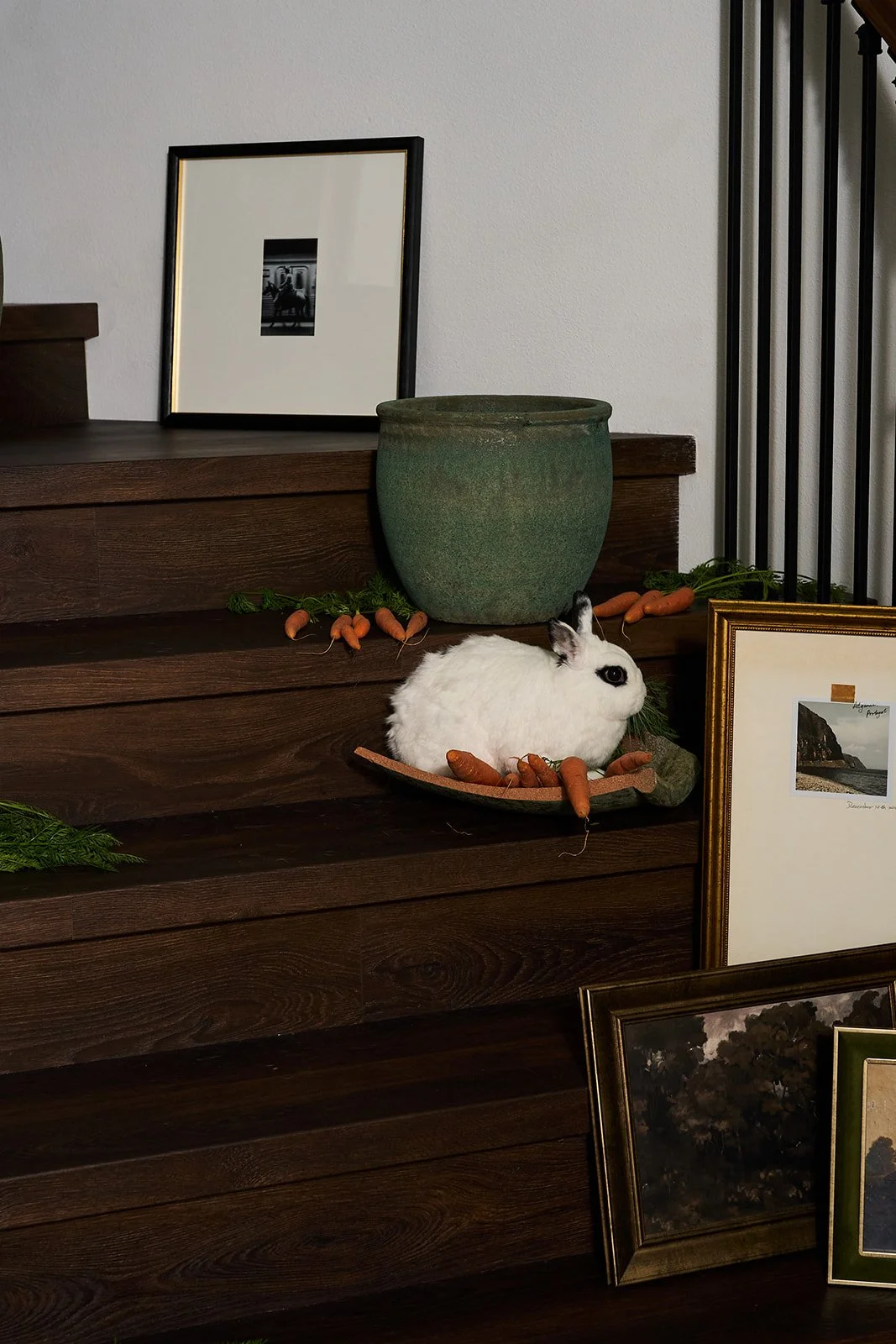 A white rabbit with black markings sits on a wood surface surrounded by orange carrots and greenery. Behind the rabbit, there is a large green vase, framed photographs, and some additional pictures leaning against a wooden surface. The scene appears 