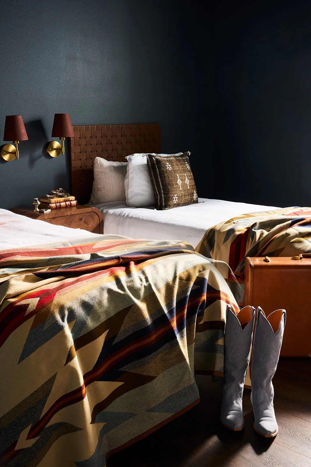 Two beds with patterned blankets in a bedroom with dark walls, vintage lamps, and cowboy boots by the foot of the bed.