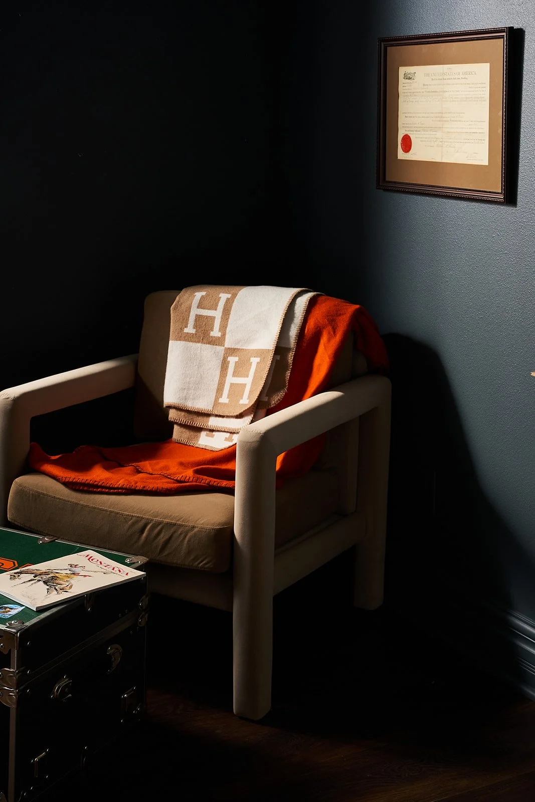 A beige armchair with a folded tan and white 'H' patterned blanket and an orange throw on it, in a dark room with navy walls. A framed document hangs on the wall above the chair, and a small black storage chest, with a magazine on top, sits in front 