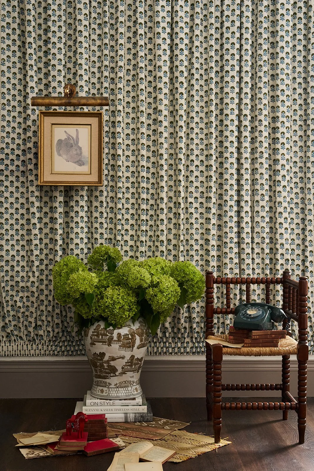 A decorated interior space featuring a floral patterned curtain, a framed rabbit illustration, a large ceramic vase with green flowers, a small wooden chair with a vintage black rotary phone, books, and decorative items on a rug.