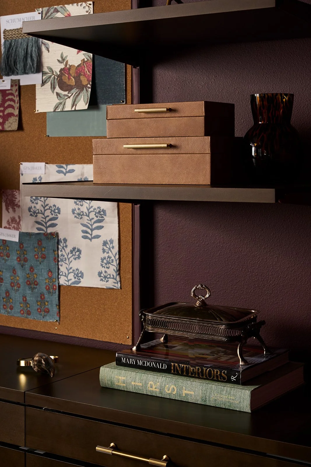 A dark shelf with two beige boxes with gold handles, a black and brown leopard-print vase, a decorative silver box on books titled 'Interiors' by Mary McDonald and 'Art' on a brown dresser, and a corkboard behind with fabric swatches and design sampl