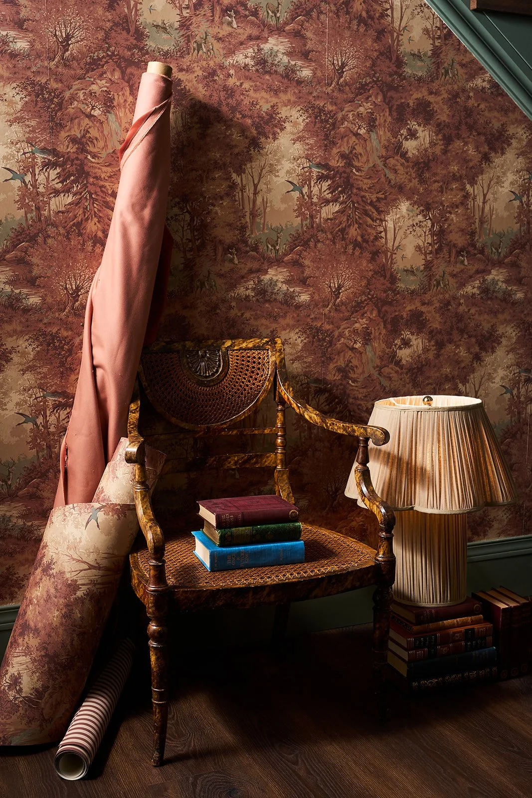 Interior room with vintage decor including a wooden armchair with a woven seat, a table lamp with a pleated shade, a stack of four colorful books on the chair, a pink drape hanging, a rolled-up wallpaper on the dark wood floor, and wallpaper with a n