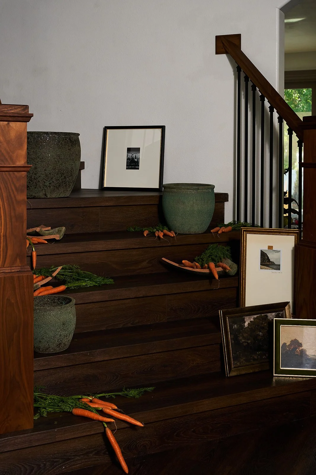 Decorative display of framed photos, ceramic pots, and real carrots arranged on dark wooden stairs in a home interior.