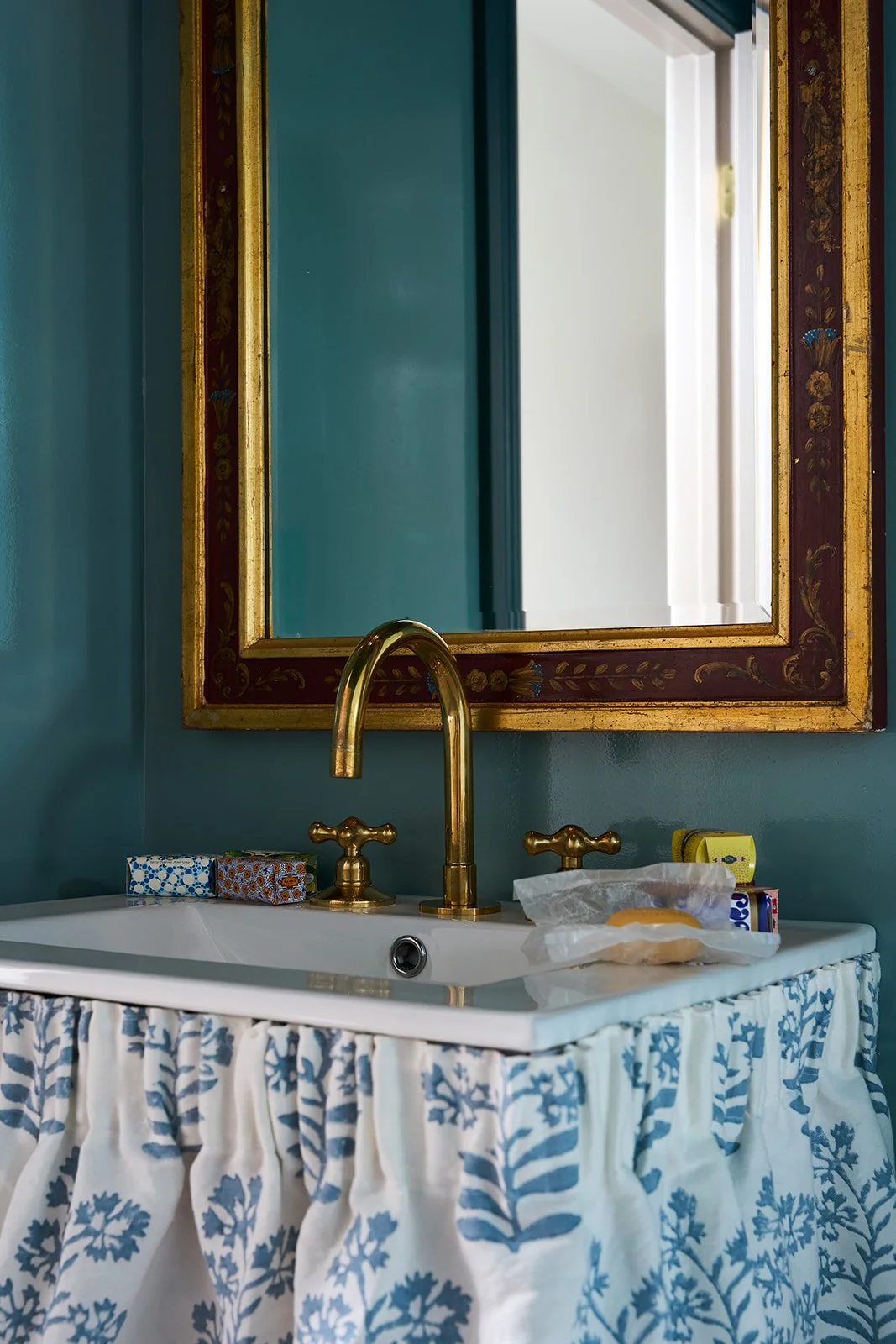 Bathroom with a white sink, gold faucet, mirror with a decorative gold and red frame, blue wall, and countertop items including a tissue box and soap.