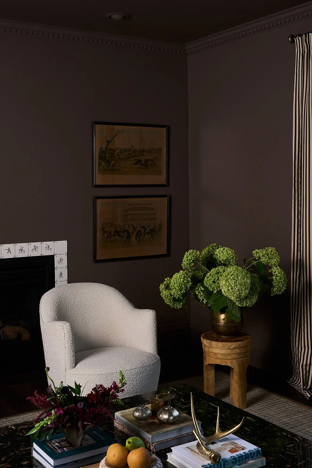 A cozy living room corner with a white textured armchair, a side table with a large green hydrangea plant, framed artwork on the wall, and a table with books, decorative objects, and fruits.
