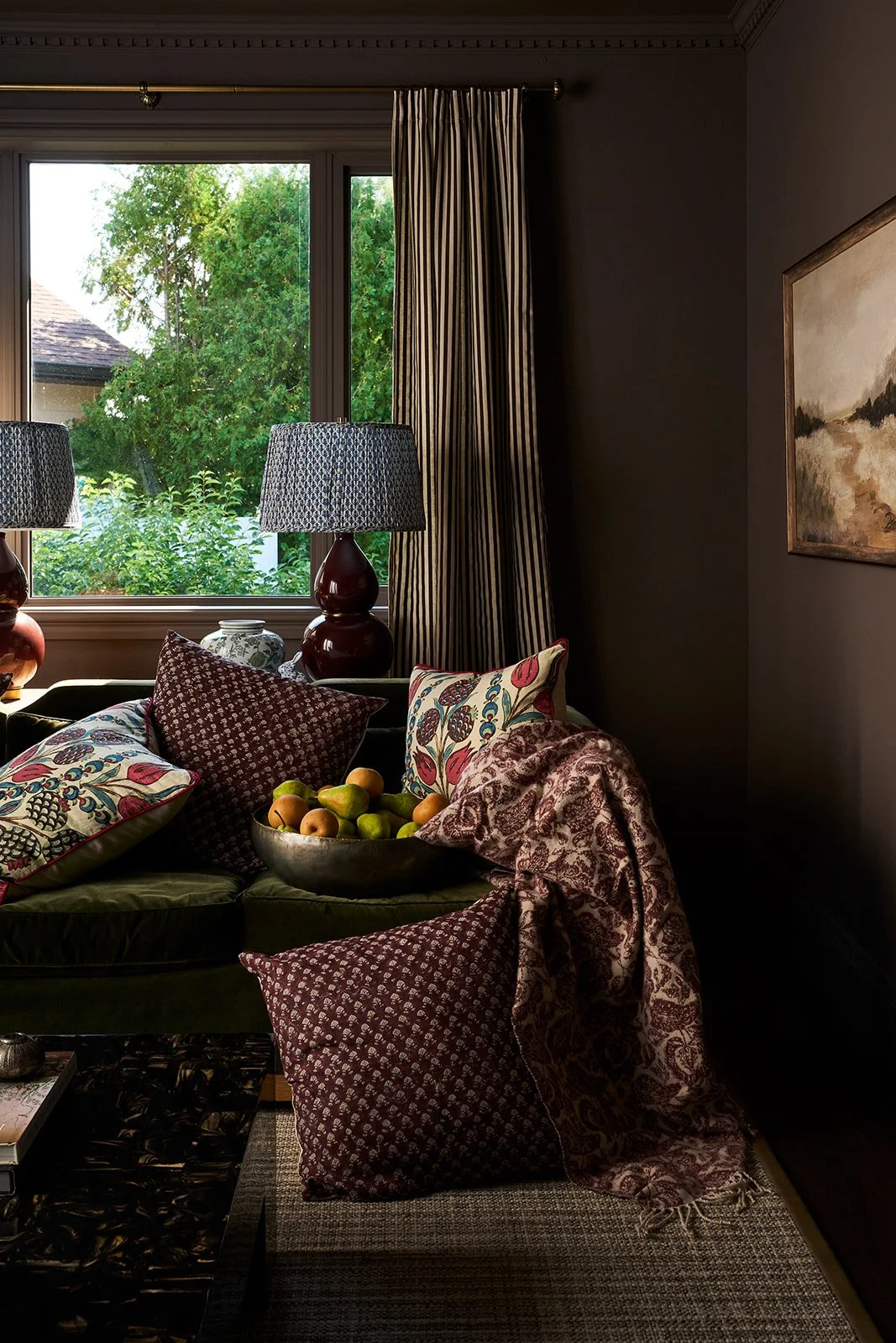 Living room with dark walls, patterned pillows and throws on a sofa, a bowl of apples and pears, a window with striped curtains, and two matching table lamps with dark red bases and patterned shades.