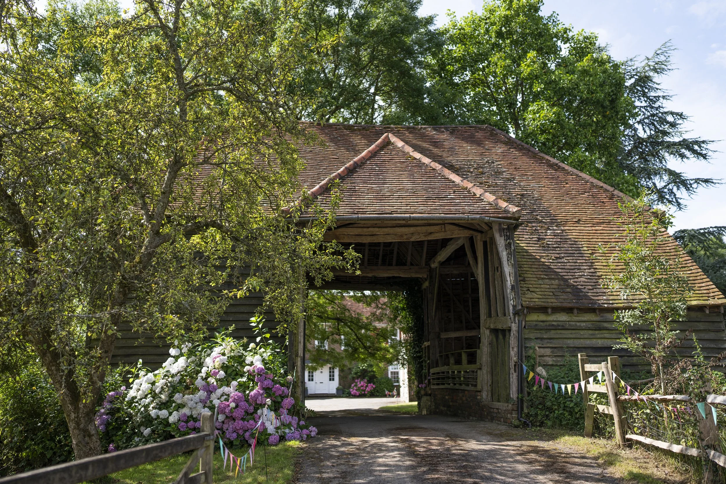 An old barn with a red-tiled roof is seen through green trees and flowering bushes, with a pathway leading towards a white building in the background. Colorful bunting flags decorate the foreground.