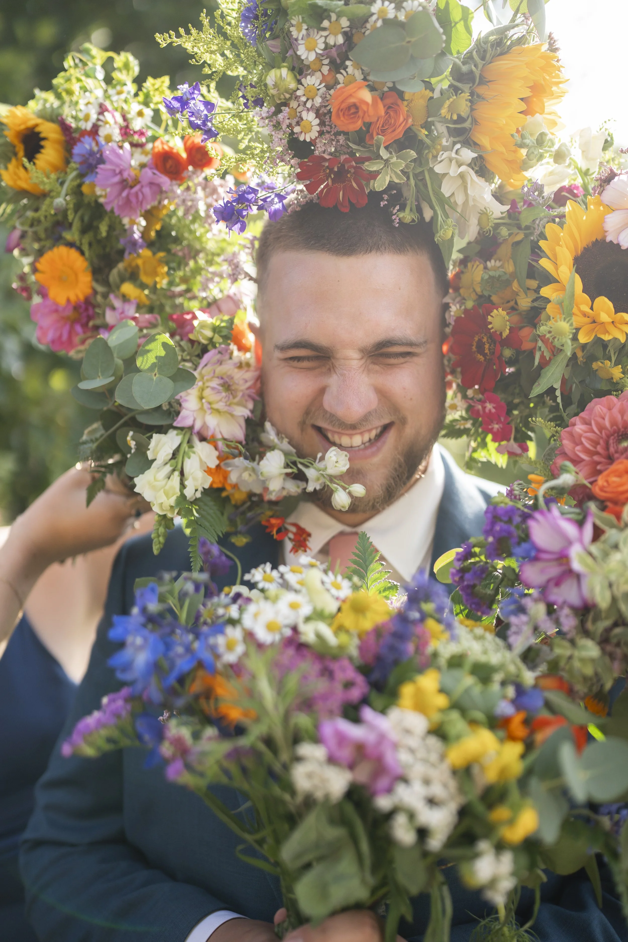 A smiling man in a suit holding a large bouquet of colorful flowers, with a floral crown on his head, outdoors on a sunny day.