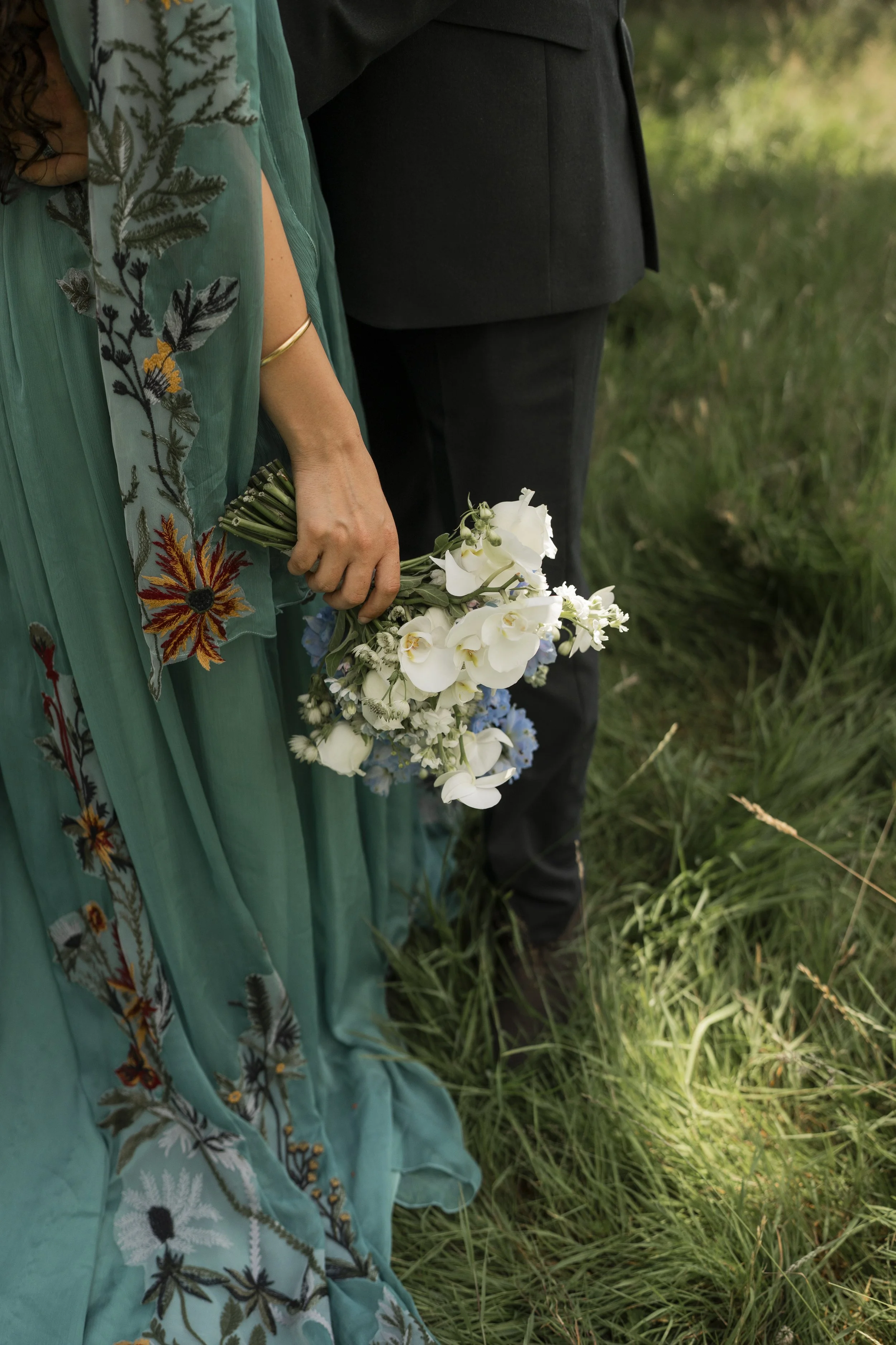 Close-up of a woman's hand holding a bouquet of white and light blue flowers, with her wearing a teal dress with floral embroidery, and a gold bracelet. Next to her, a man is wearing a black suit, and they are standing on green grass.