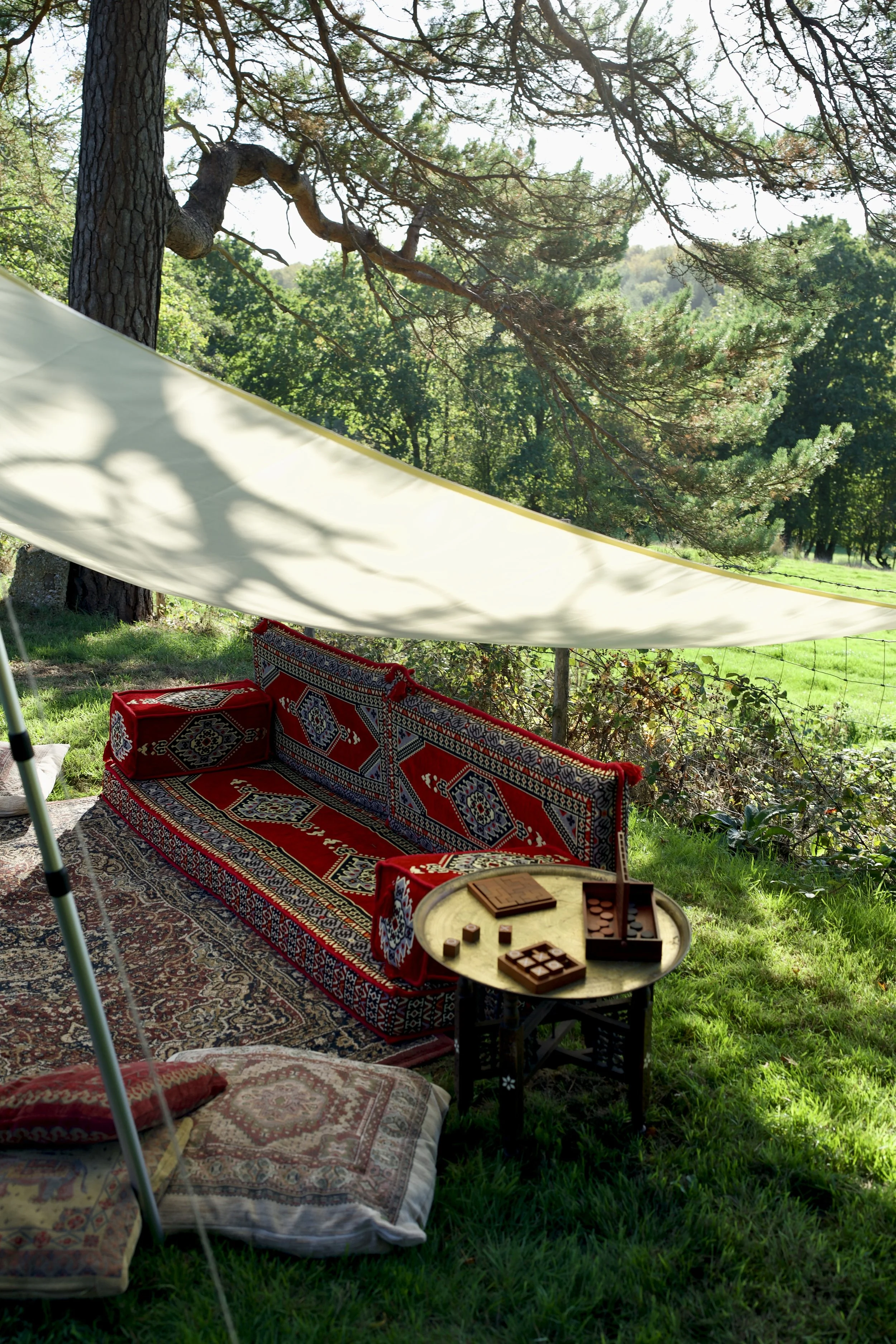 Outdoor seating area with patterned cushions and rugs under a large tree, with a chessboard and game pieces on a small round table, and a cozy shaded space with a white canopy.