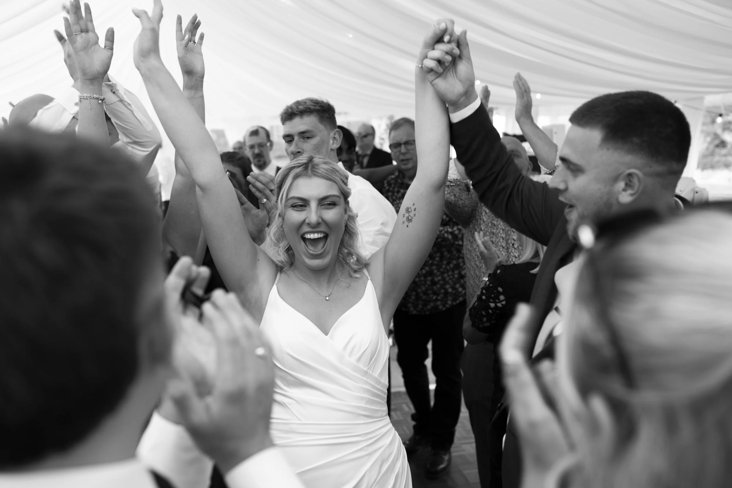 A woman in a white dress dancing and smiling, surrounded by people at a celebration or wedding reception, raising her arms in joy.