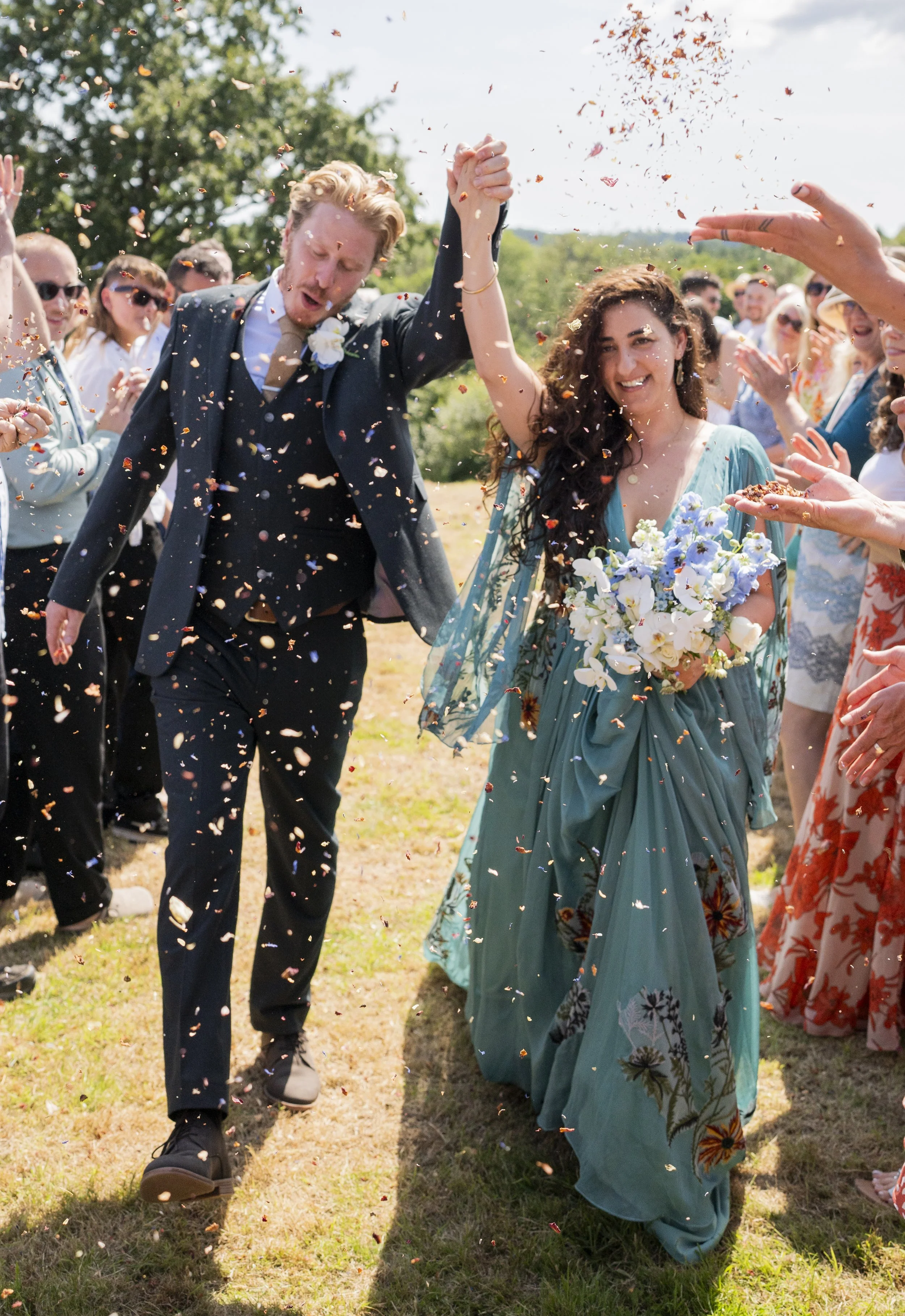 A wedding celebration outdoors with a bride and groom holding hands and walking on a grassy area, surrounded by guests throwing confetti. The groom has light hair and wears a dark suit, while the bride has dark wavy hair and wears a flowing blue dress and holds a bouquet of flowers. The scene is sunny with a backdrop of trees and a blue sky.