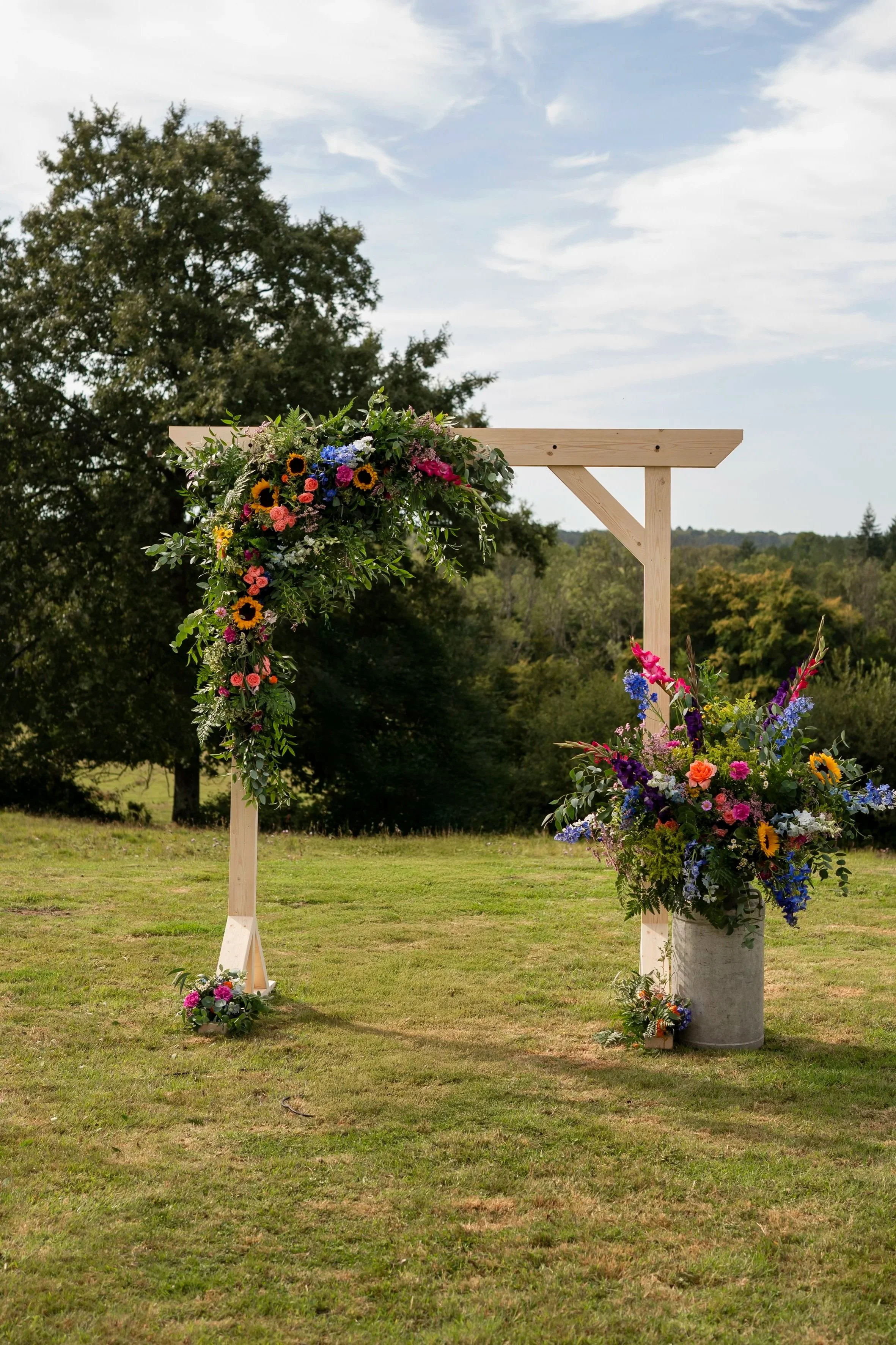 A wooden outdoor wedding altar decorated with colorful flowers, set on a grassy field with trees and a partly cloudy sky in the background.