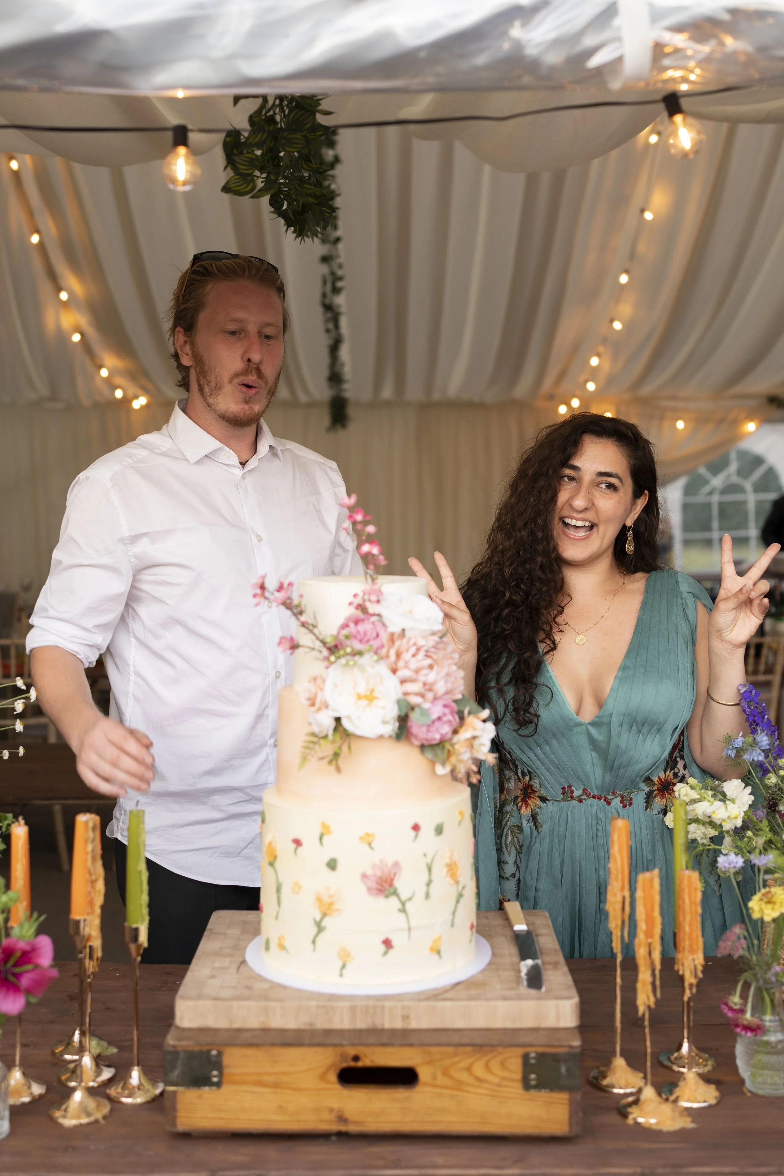 A group of friends celebrating with a layered floral cake at a party, decorated with flowers and surrounded by candles and string lights.