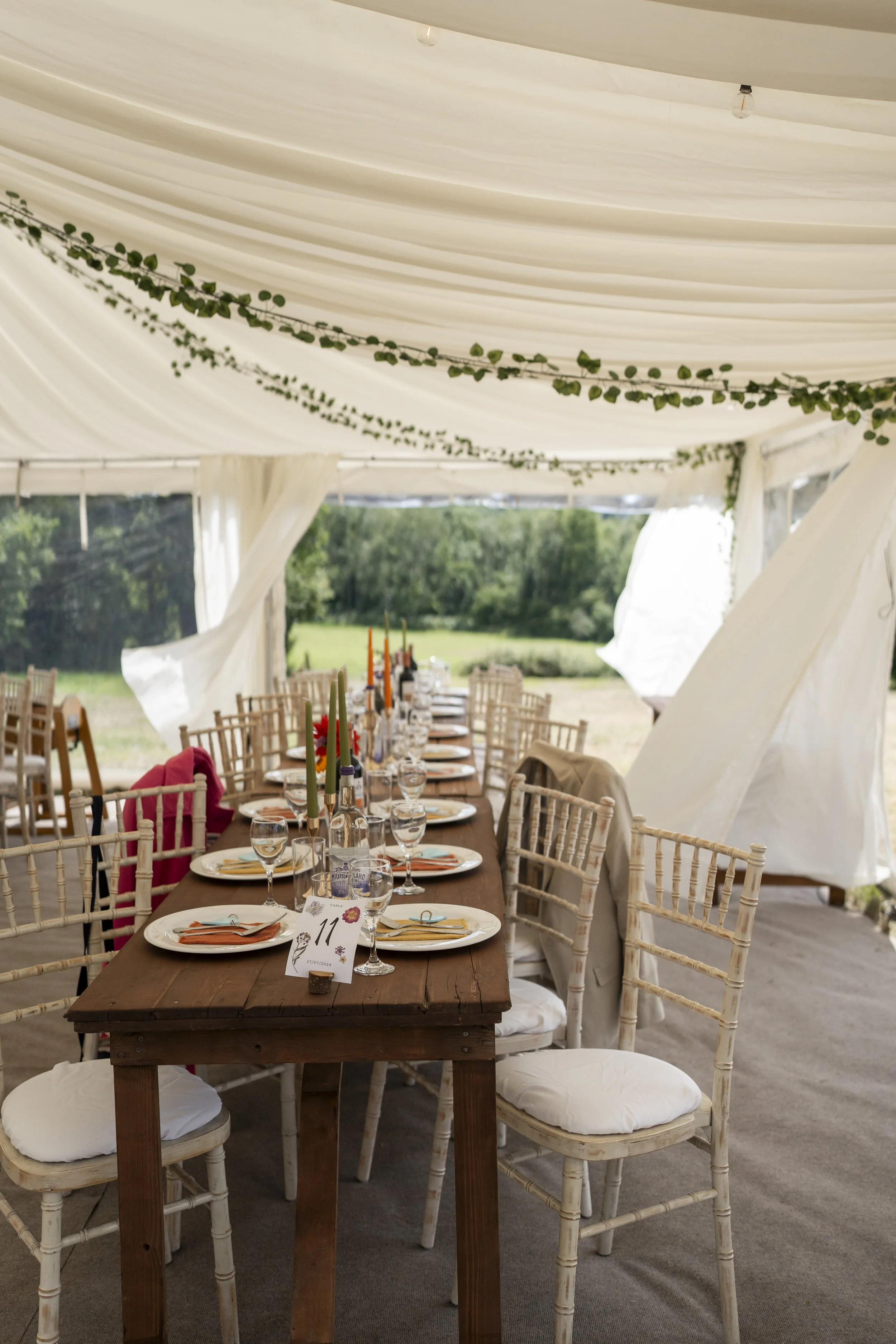A long wooden dining table set for a meal under a large white tent outdoors, with chairs around it, tableware, and numbered place cards, with greenery visible in the background.
