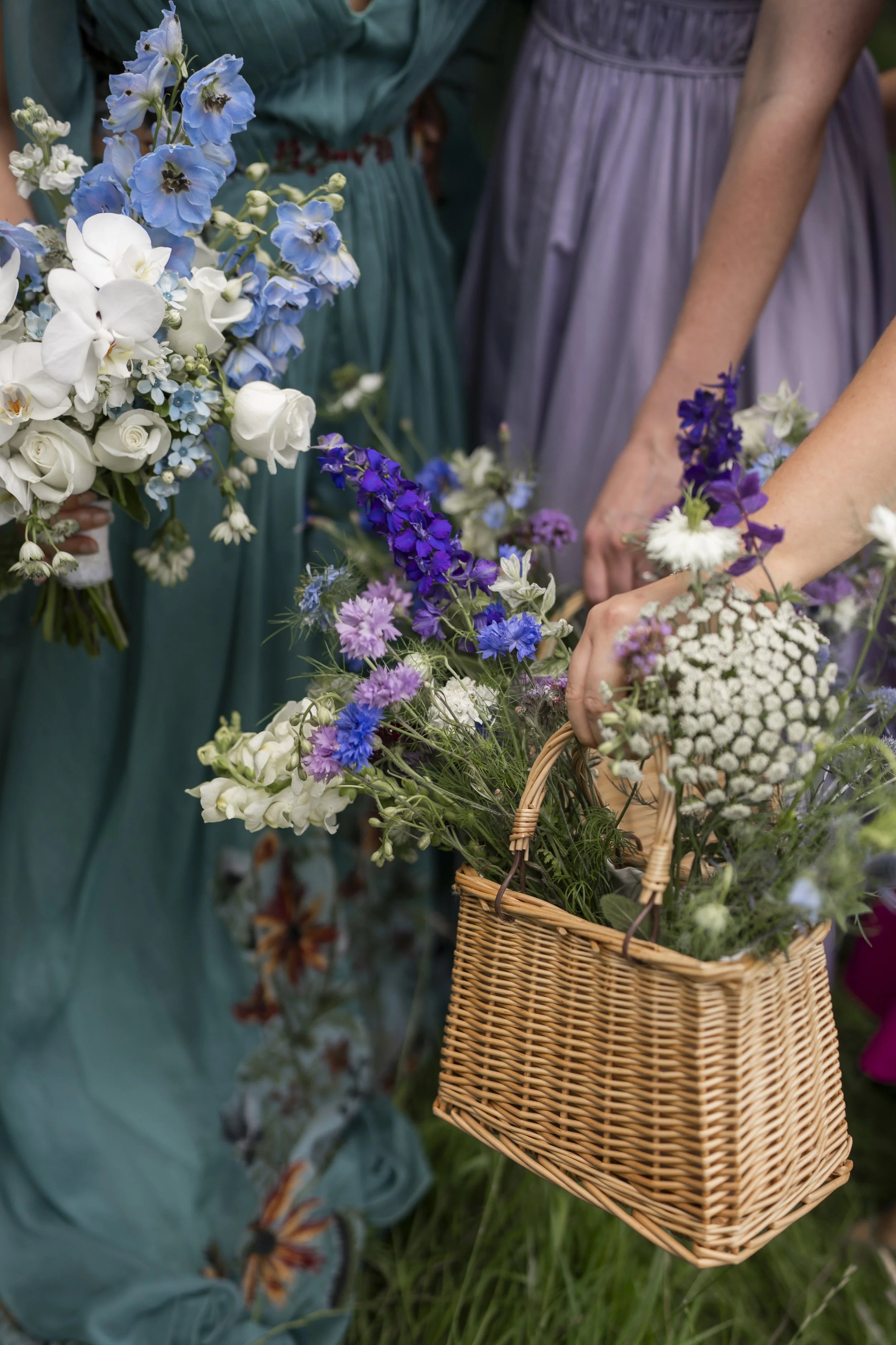Close-up of women holding a wicker basket filled with various colorful flowers, with one woman in a purple dress and another in a green dress in the background.