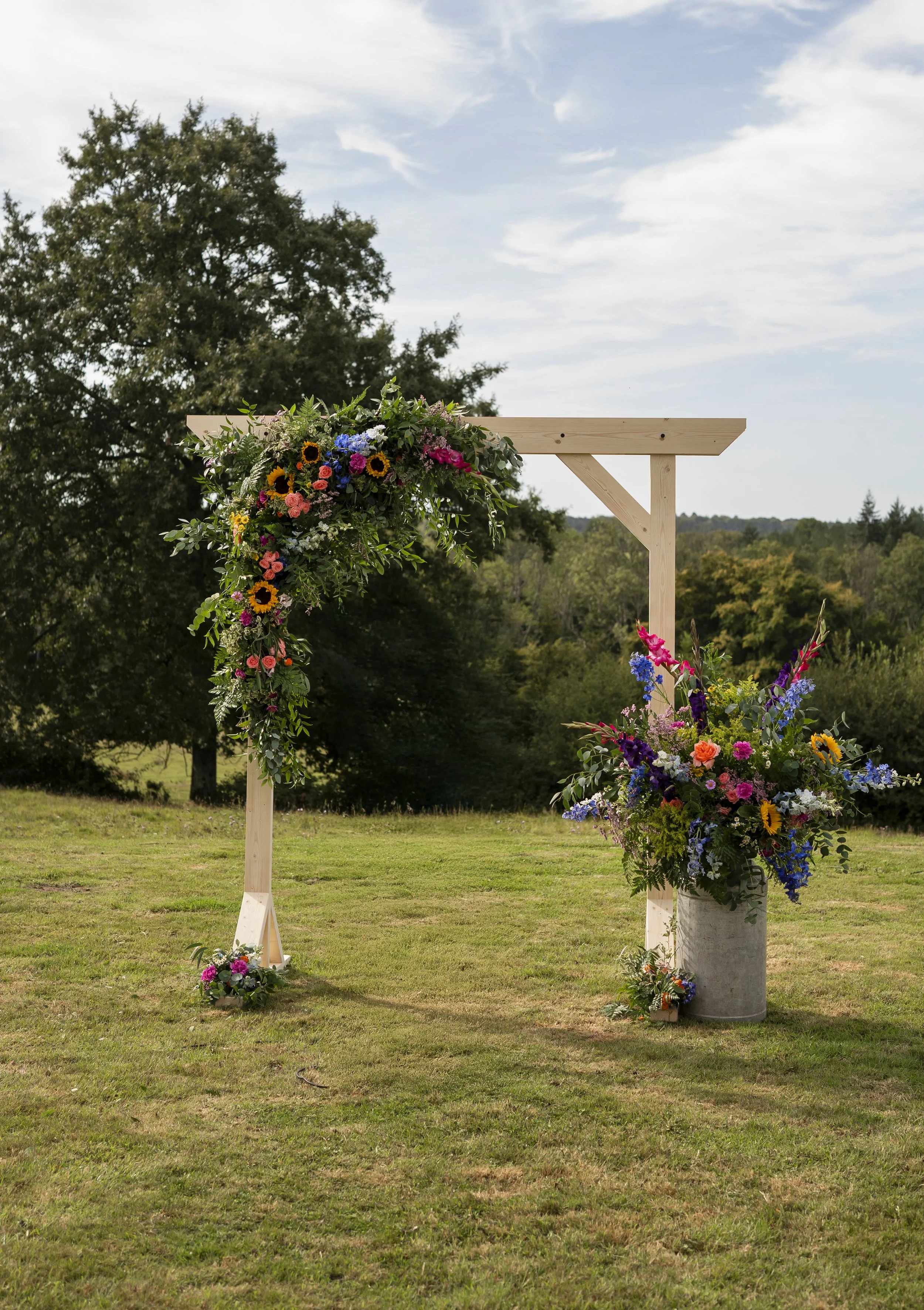 A wedding arch decorated with colorful flowers, set outdoors on a grassy field with trees and a partly cloudy sky in the background.