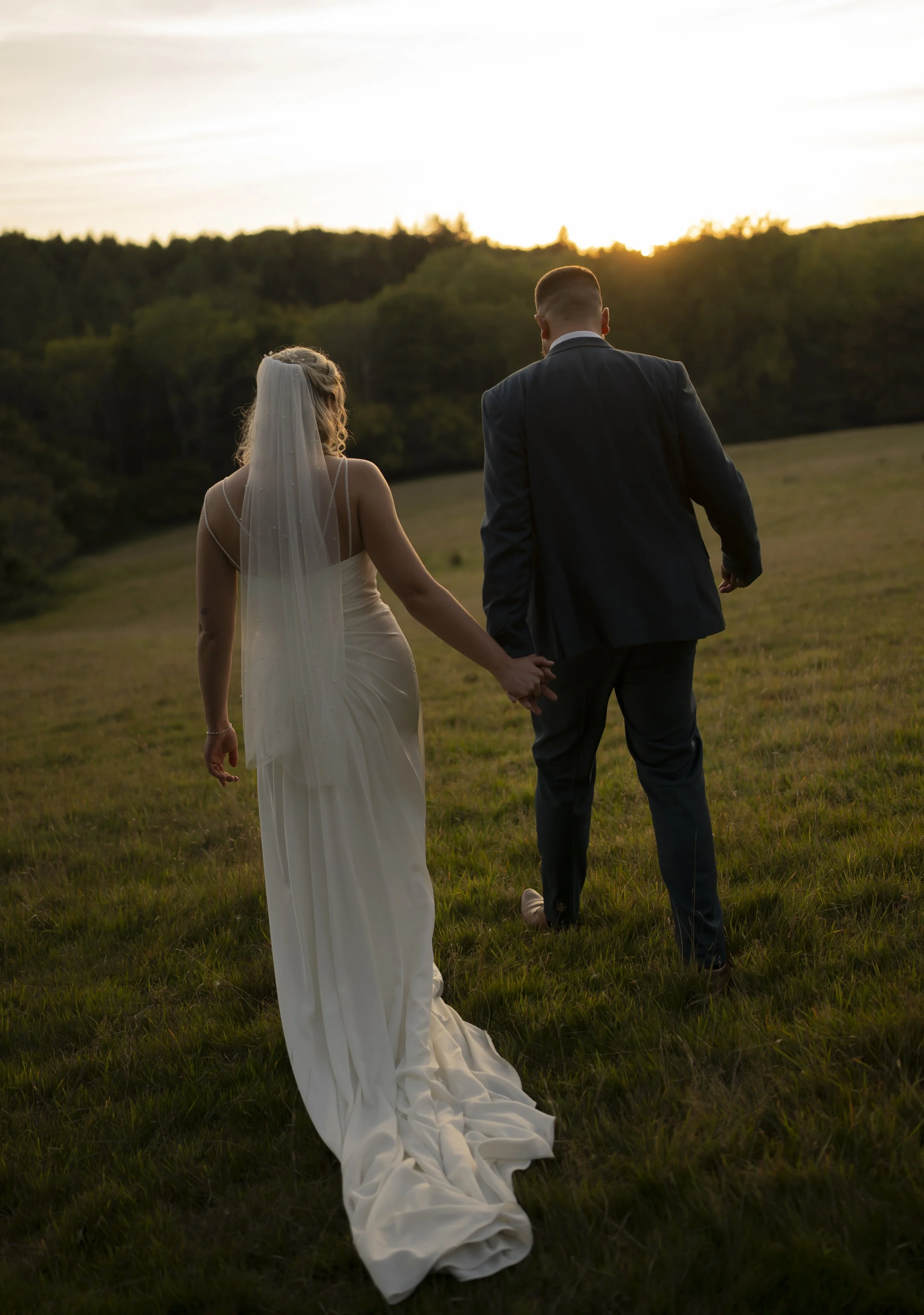 A bride and groom holding hands walk through a grassy field at sunset, with trees and a glowing sky in the background.