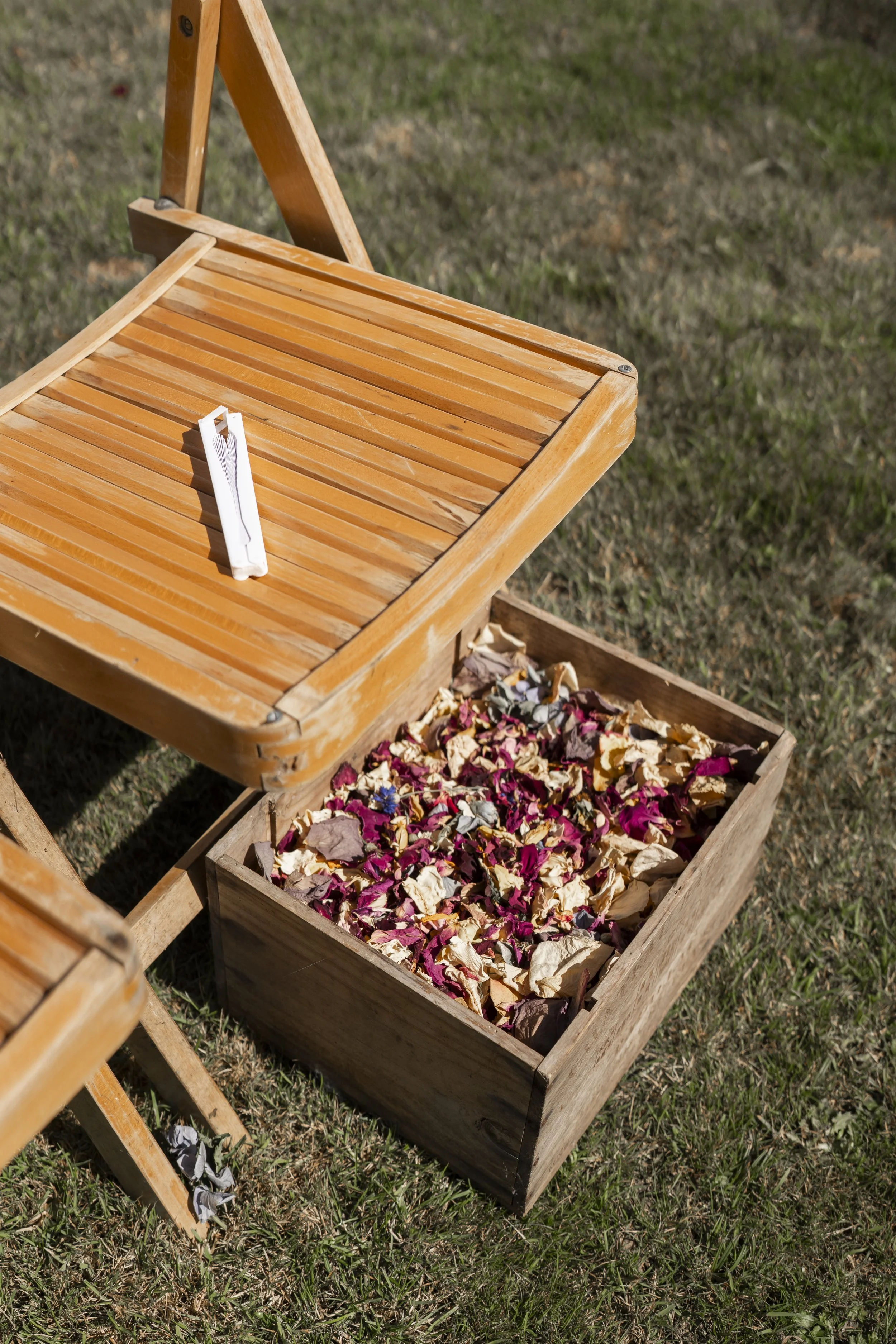 Wooden folding chair and wooden box filled with dried flower petals outdoors on grass.