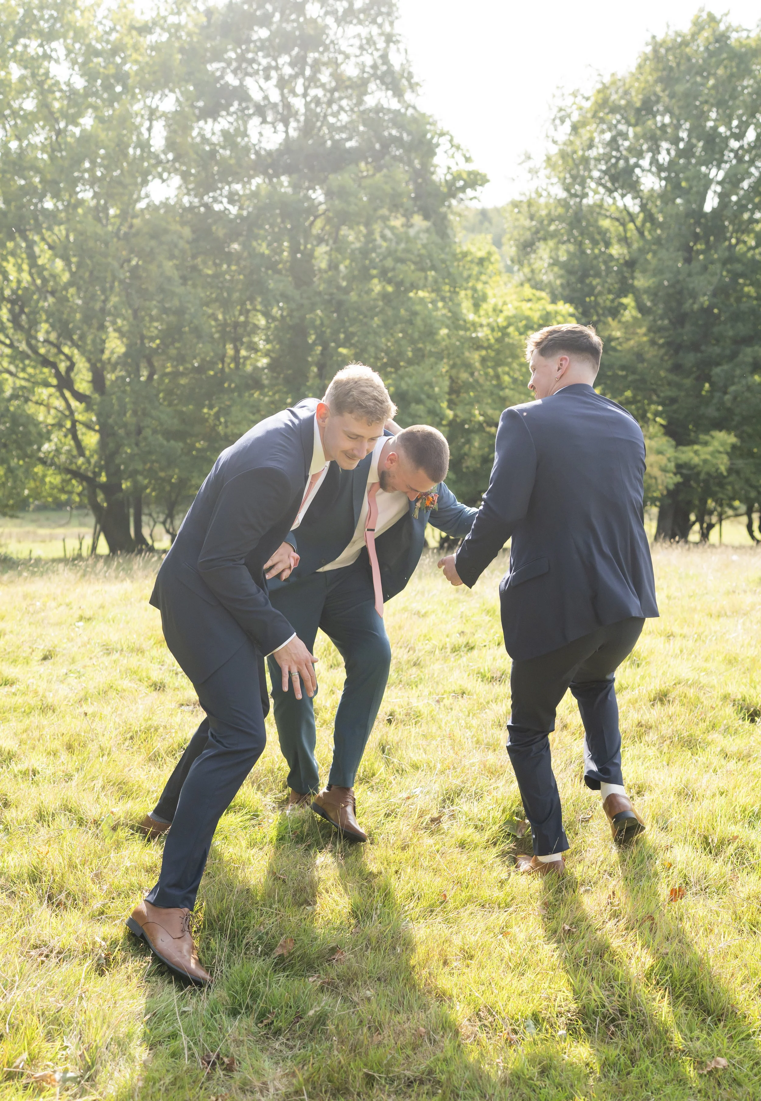 Four men in suits laughing and bending forward outdoors on grass with trees in the background.