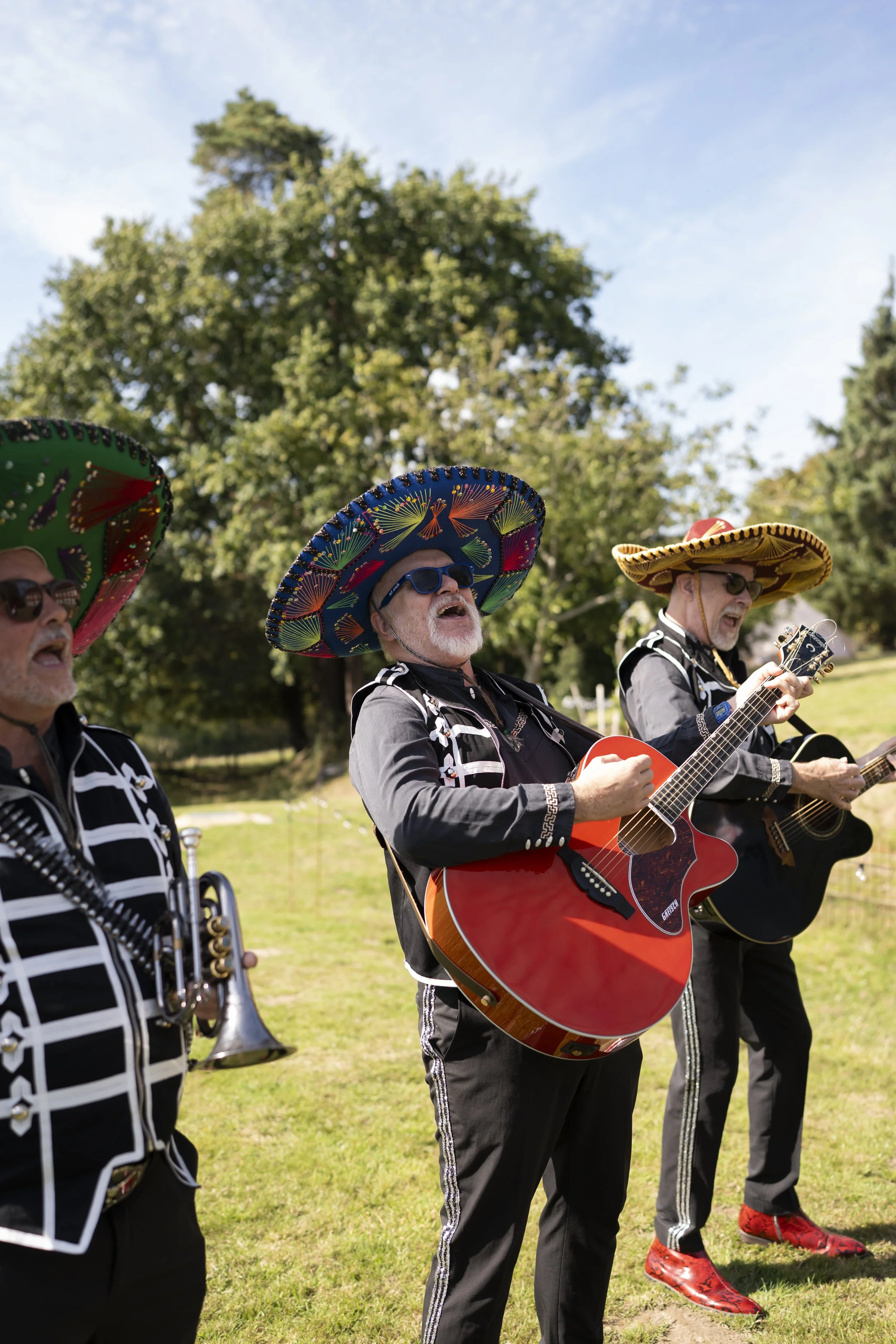 Mexican musicians wearing sombreros and traditional clothing playing guitars and a trumpet in a park with green trees and blue sky.