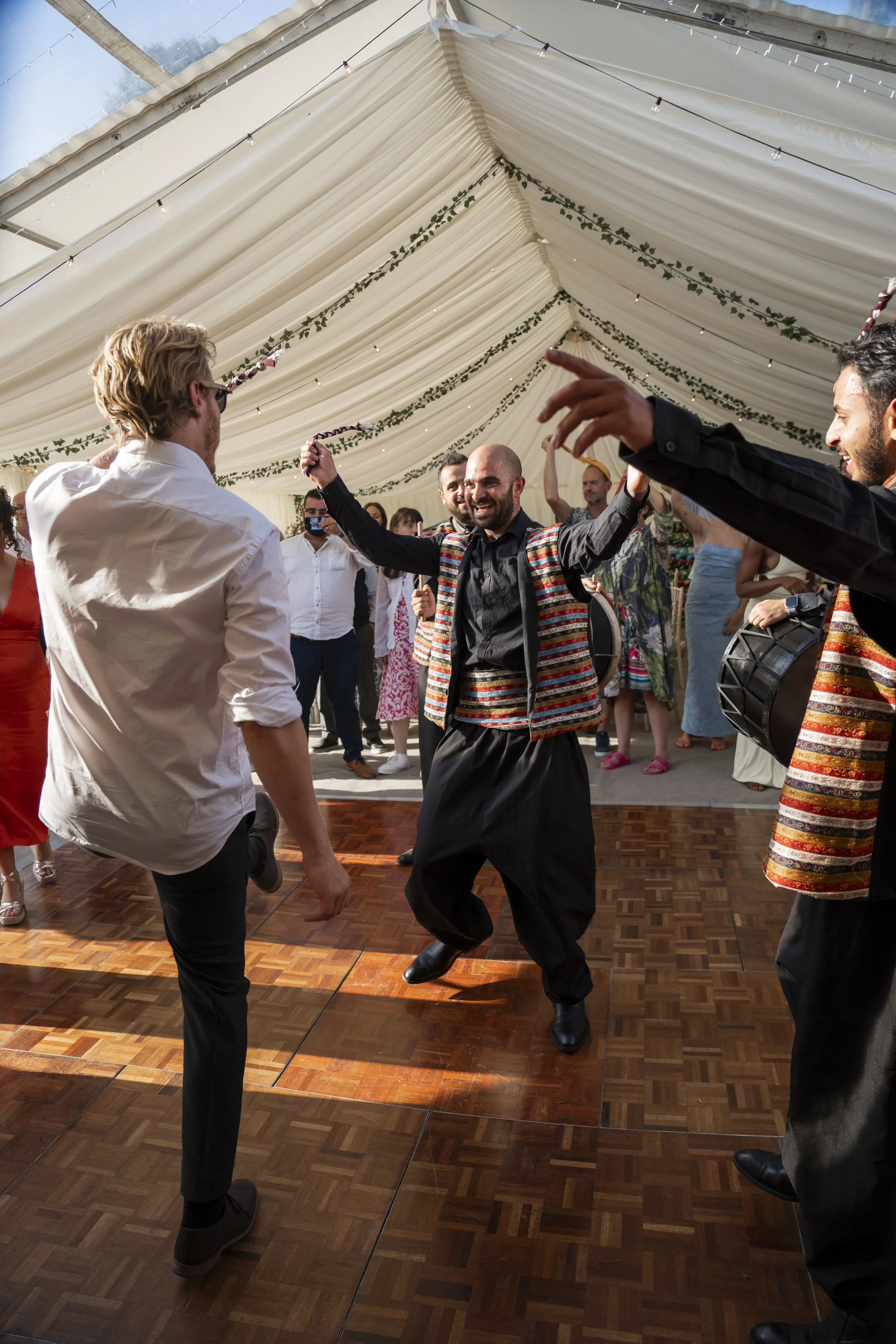 People dancing at a celebration under a decorated tent with string lights and greenery, with a wooden dance floor and a lively atmosphere.