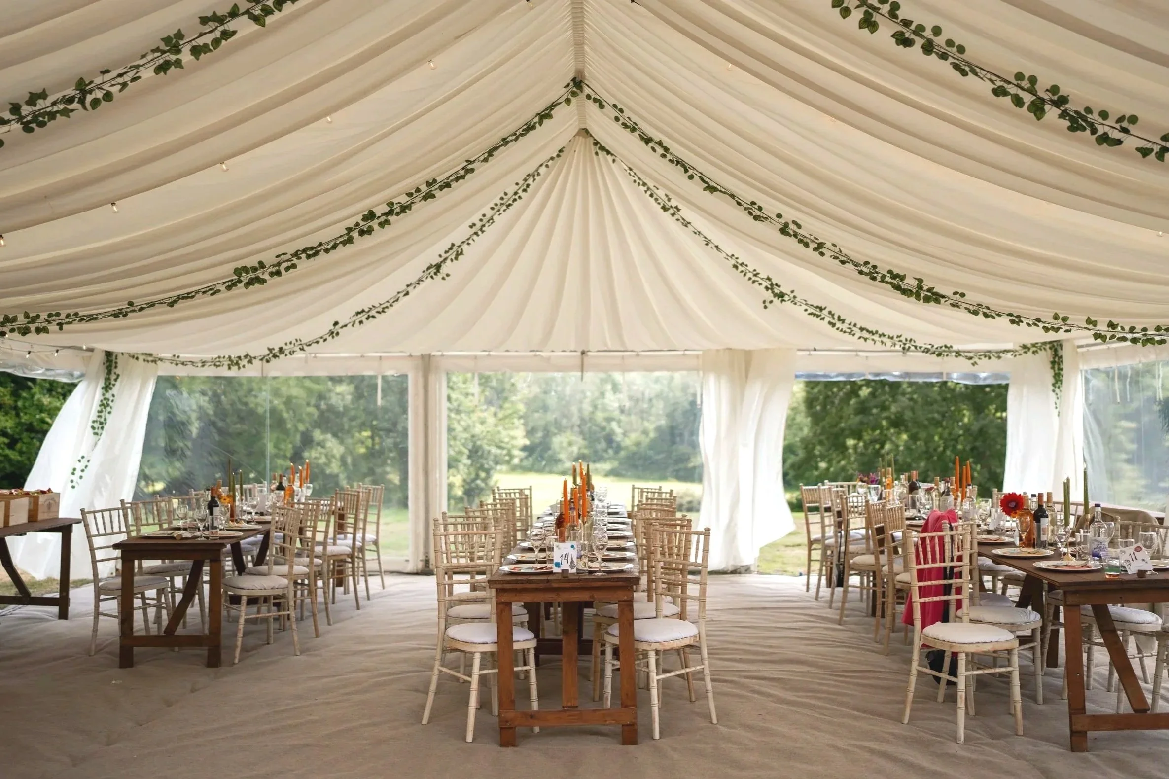Wedding reception setup under a large tent with white draped ceiling and green vine decorations. Round and rectangular tables are arranged with chairs, table settings, candles, and centerpieces. Natural scenery visible through open sides of the tent.