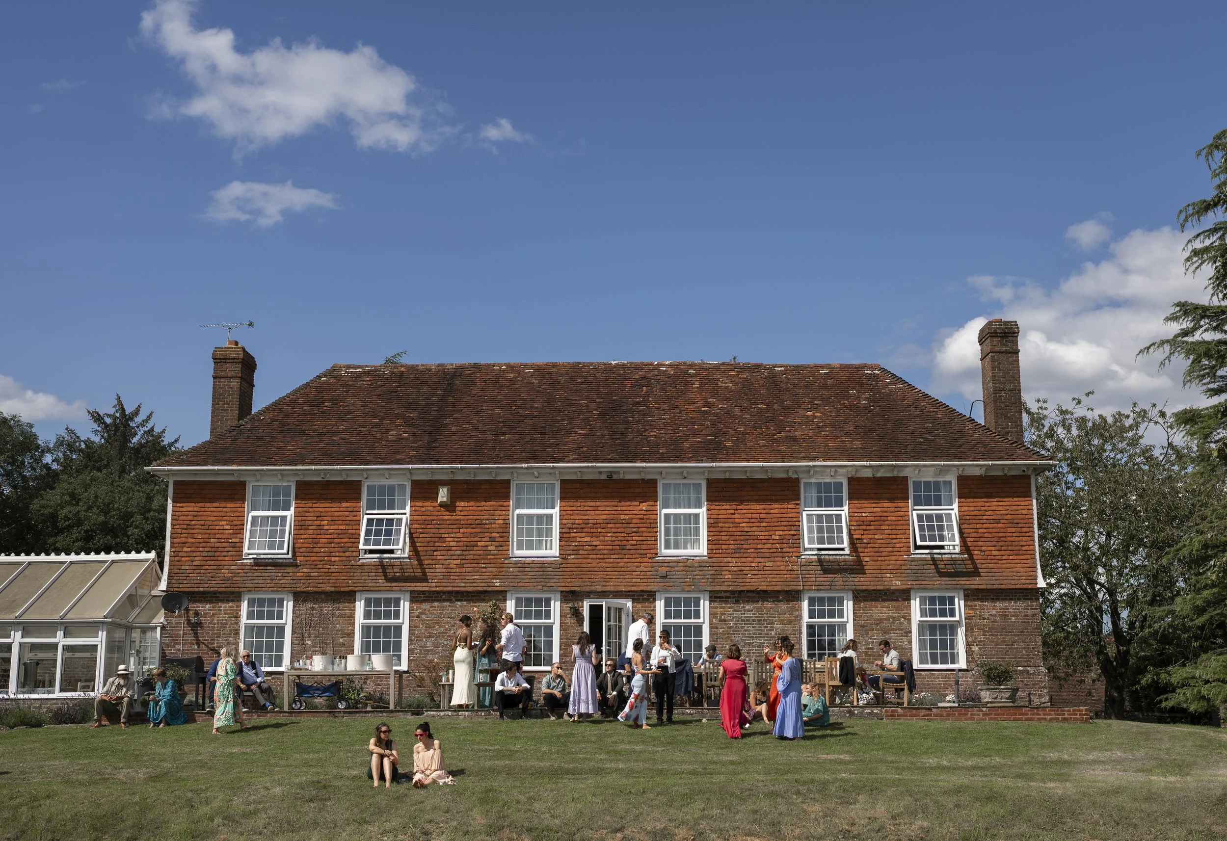 People gather on a lawn in front of a brick house with a pitched roof, attending an outdoor event on a sunny day with a blue sky and clouds.