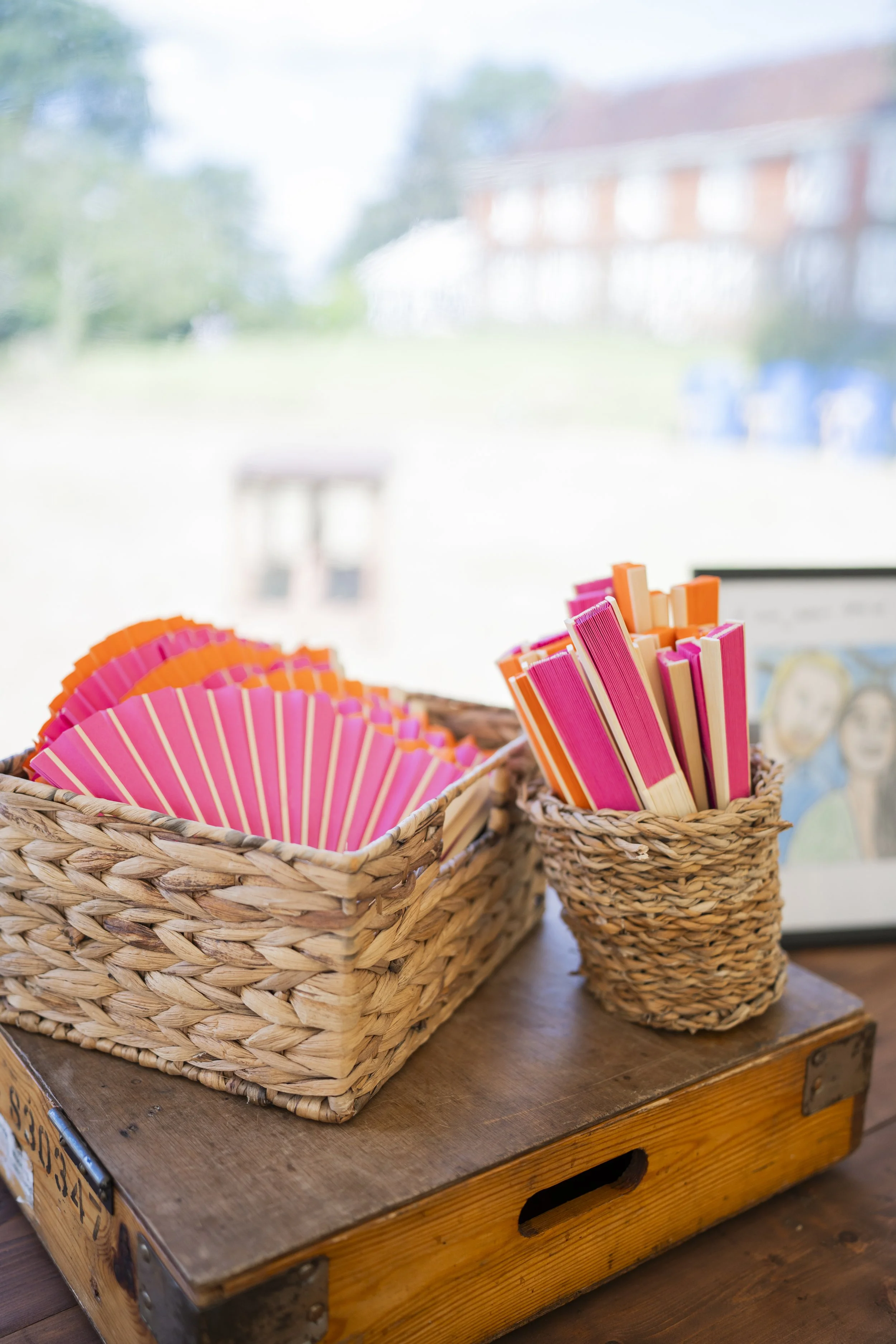 Wicker basket filled with pink and orange paper fans, and a separate smaller wicker container with colorful folded paper sticks, on a wooden surface.