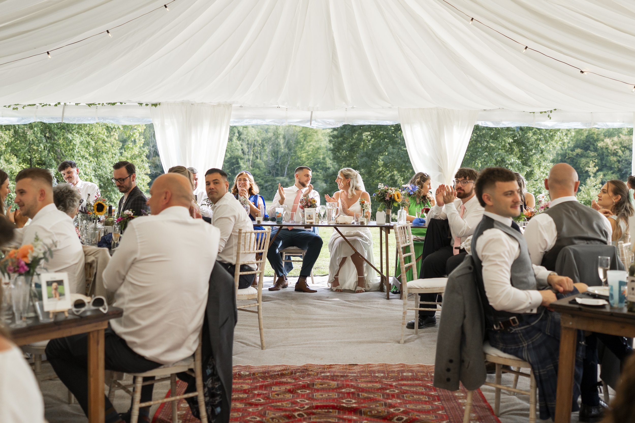 People seated at tables inside a large white tent at an outdoor wedding reception, with green trees in the background.