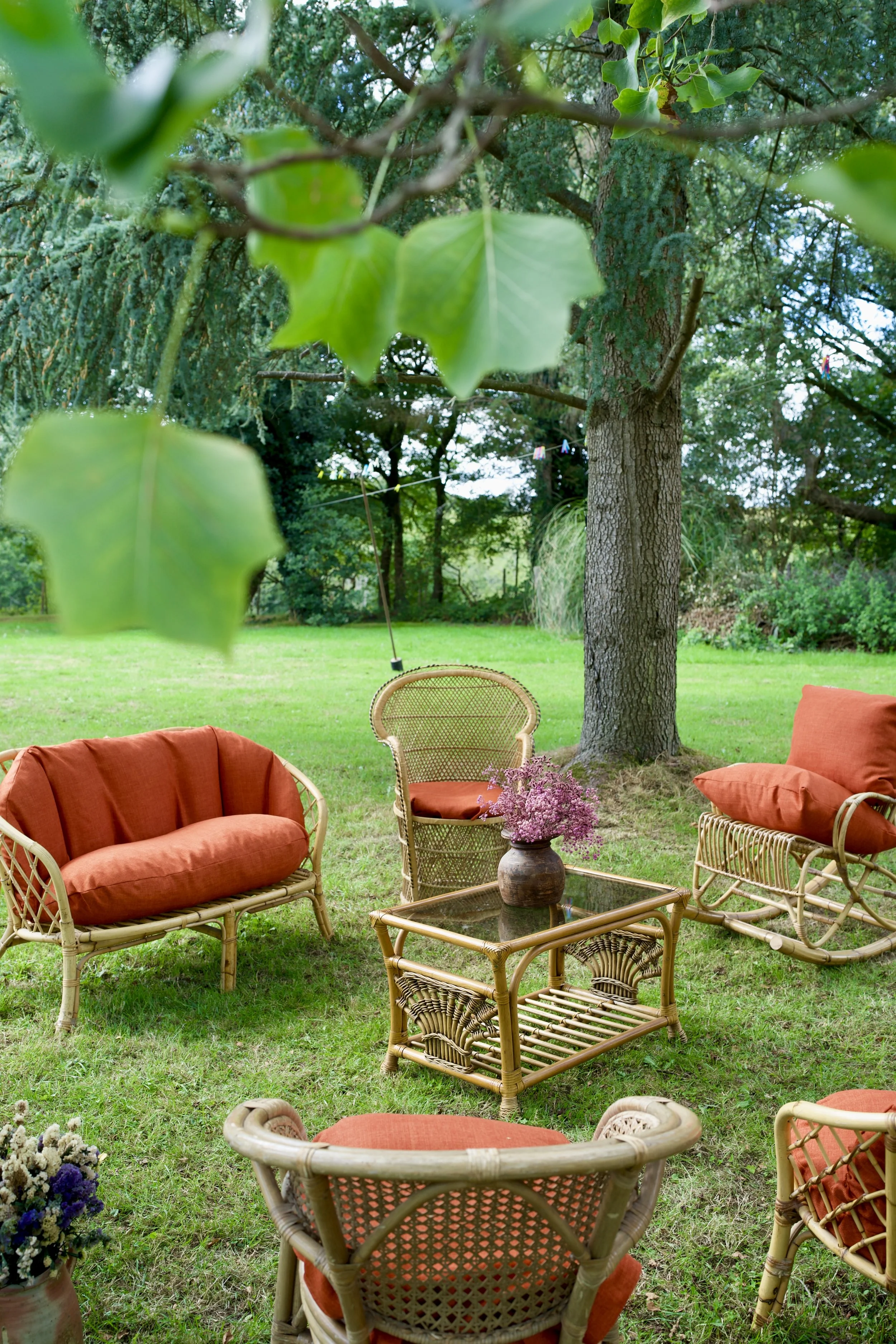Outdoor patio area with wicker chairs and cushions, a glass-top table with a vase of purple flowers, set in a garden with green grass, trees, and foliage.