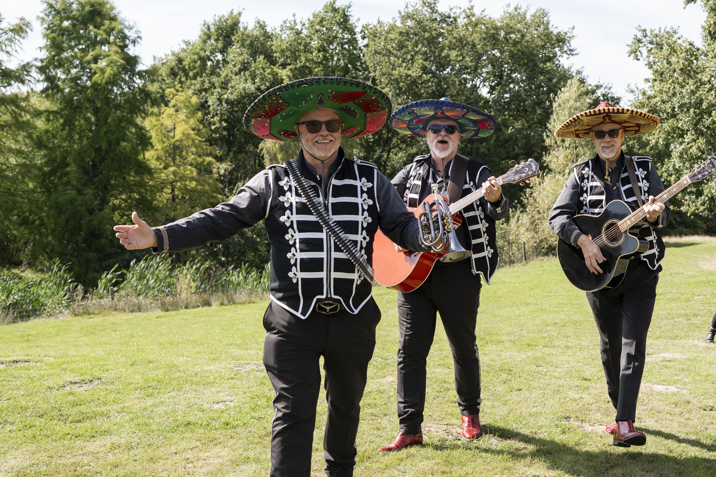 Three men wearing traditional Mexican mariachi outfits and large sombreros, singing and playing guitars outdoors on a sunny day with green trees in the background.
