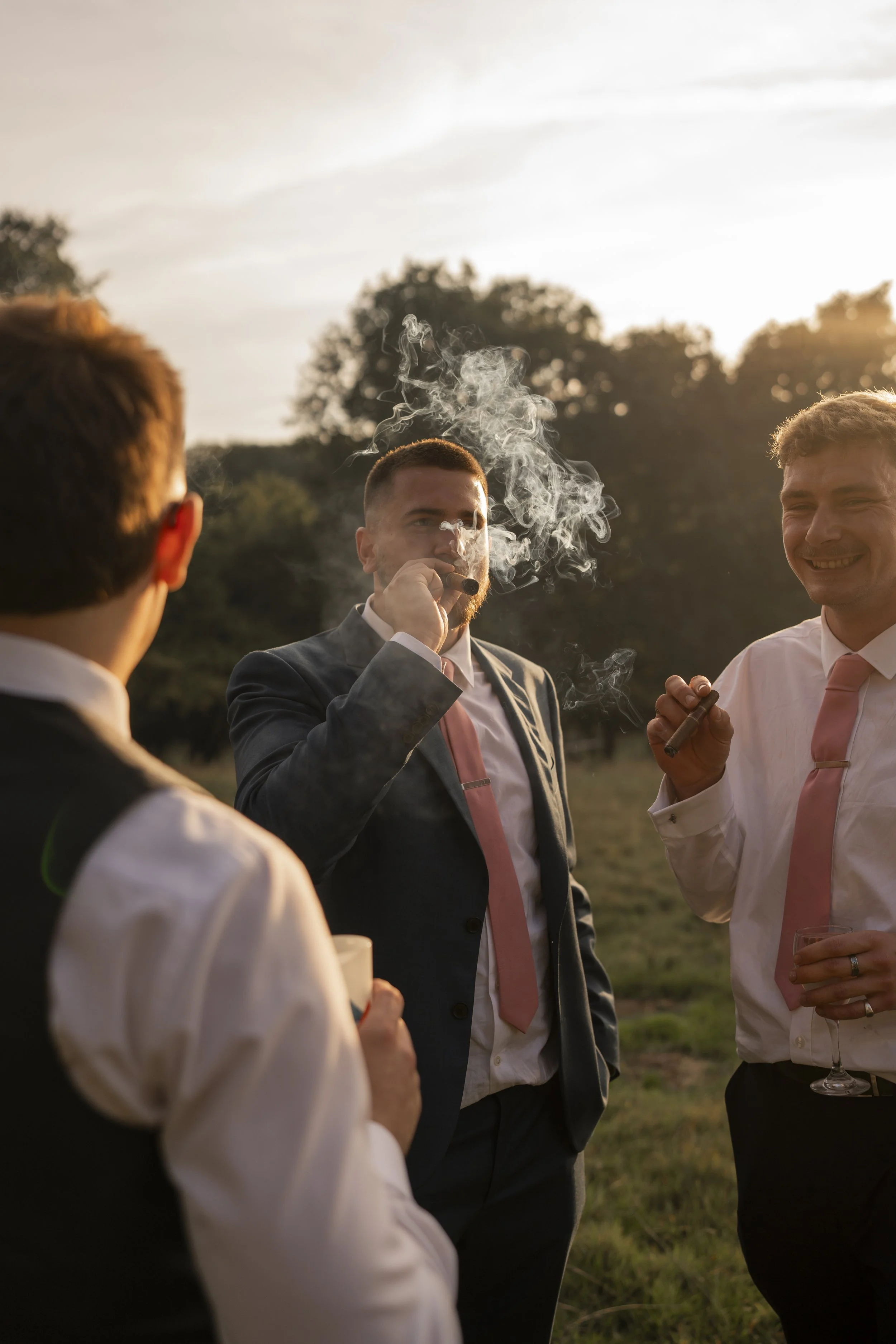 Group of men dressed in suits and shirts, some with ties, socializing outdoors during sunset, with one man smoking a cigar and others holding drinks.