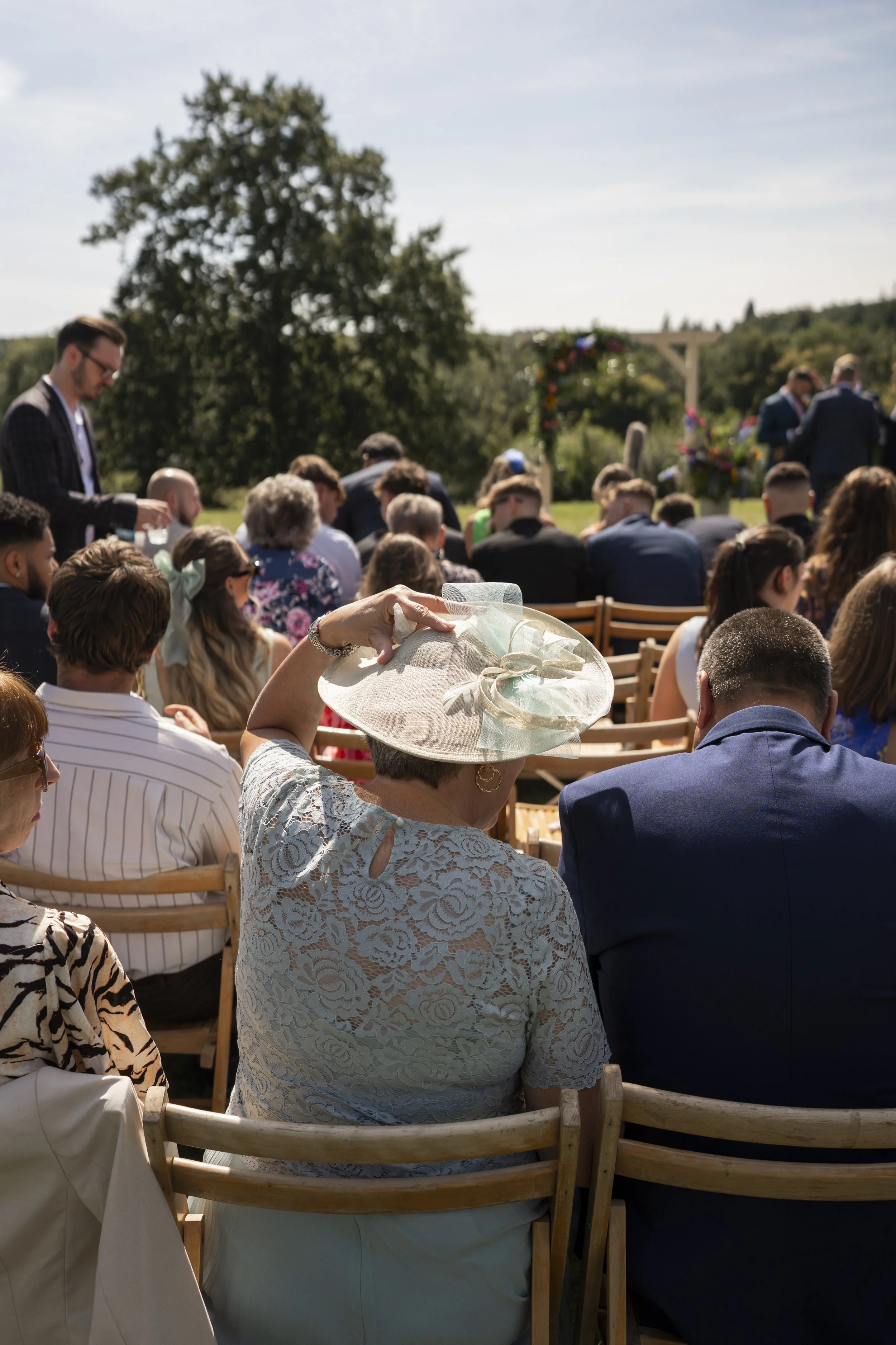 People attending an outdoor wedding ceremony, seated on wooden chairs, with floral decorations and a large tree in the background.