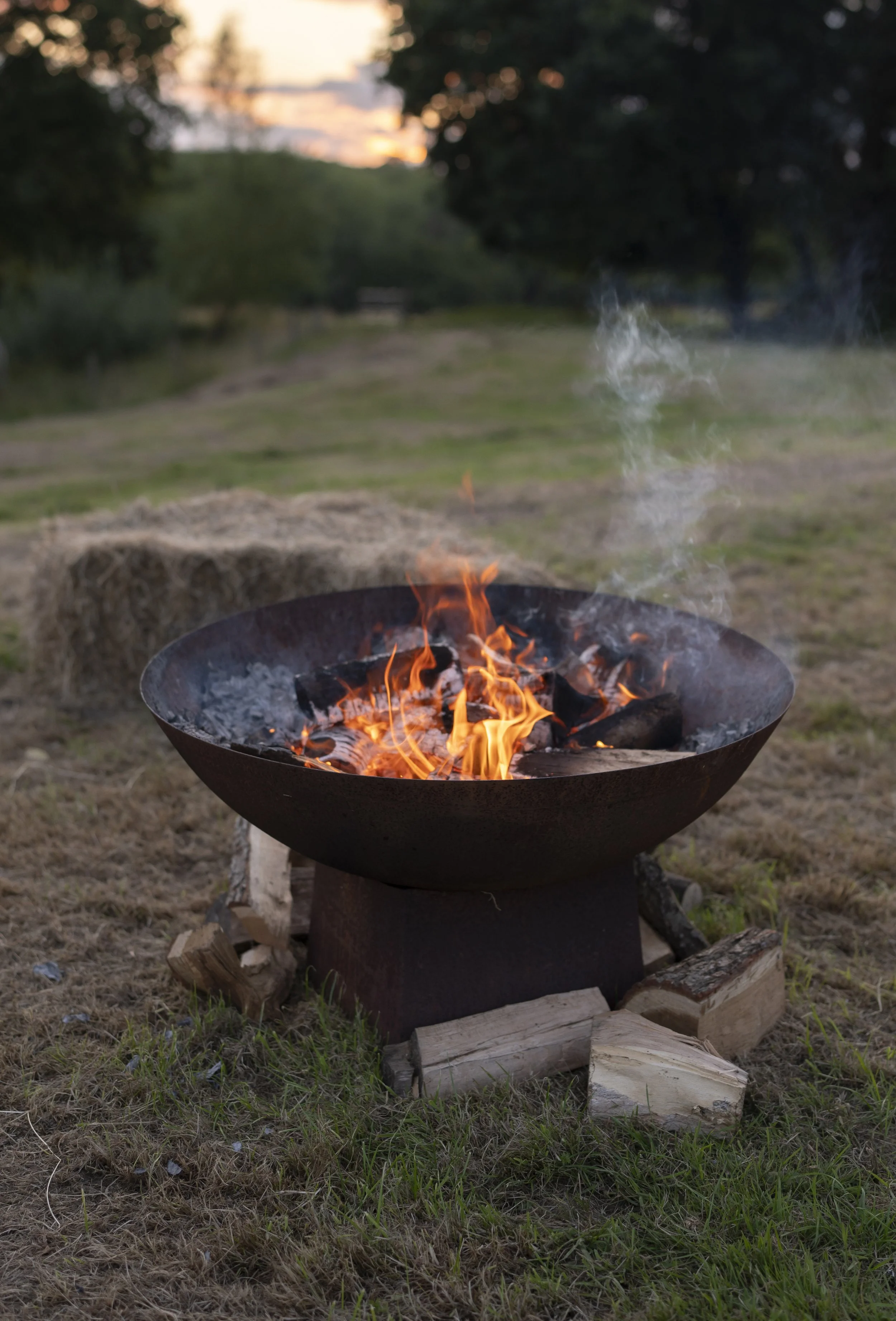 A metal fire pit with a burning fire, smoke rising, situated outdoors on a grassy area with trees and a sunset in the background.