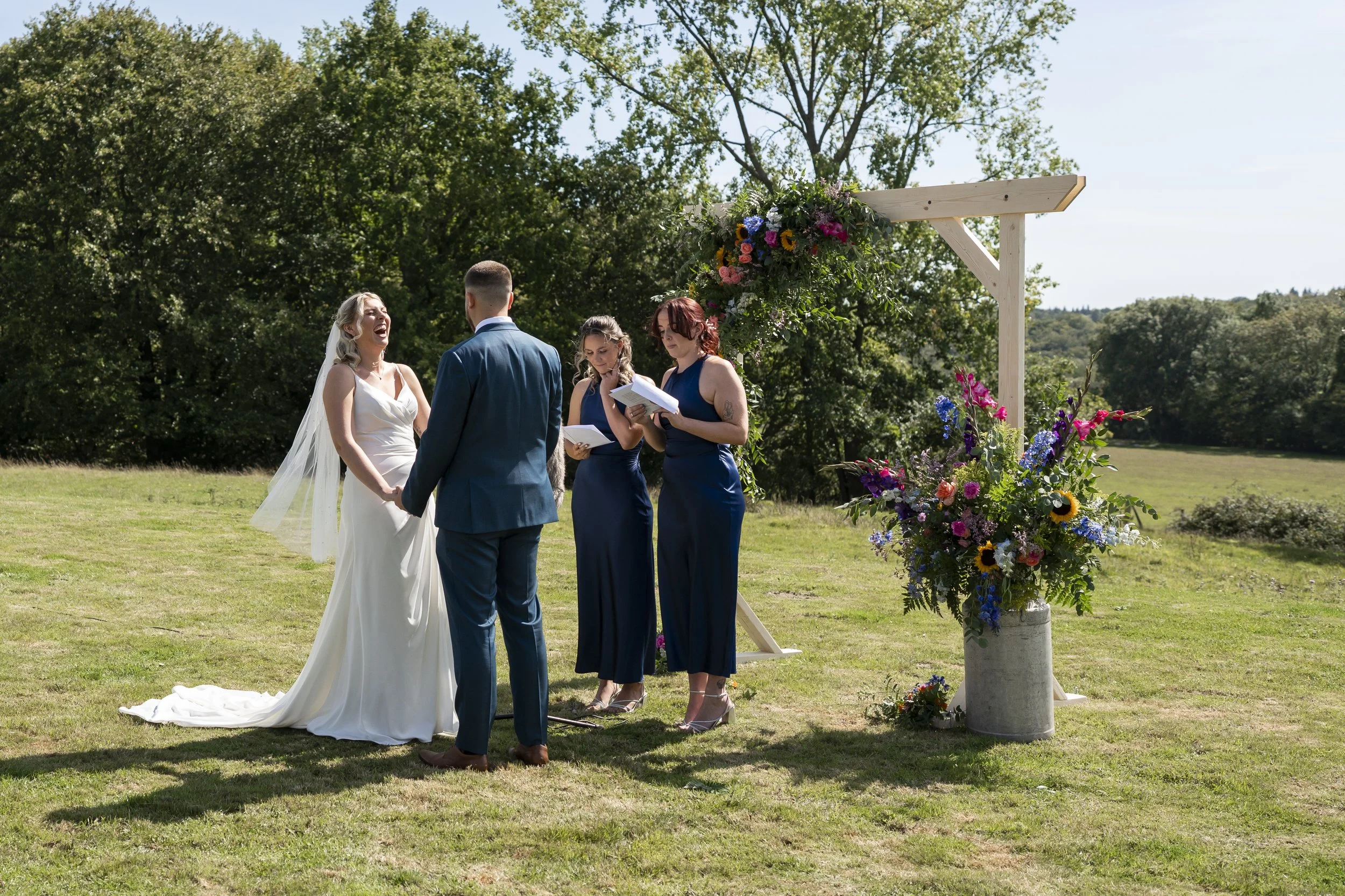 A couple getting married outdoors with an officiant and two bridesmaids, standing under a decorated wooden arch with colorful flowers on a grassy field surrounded by trees.