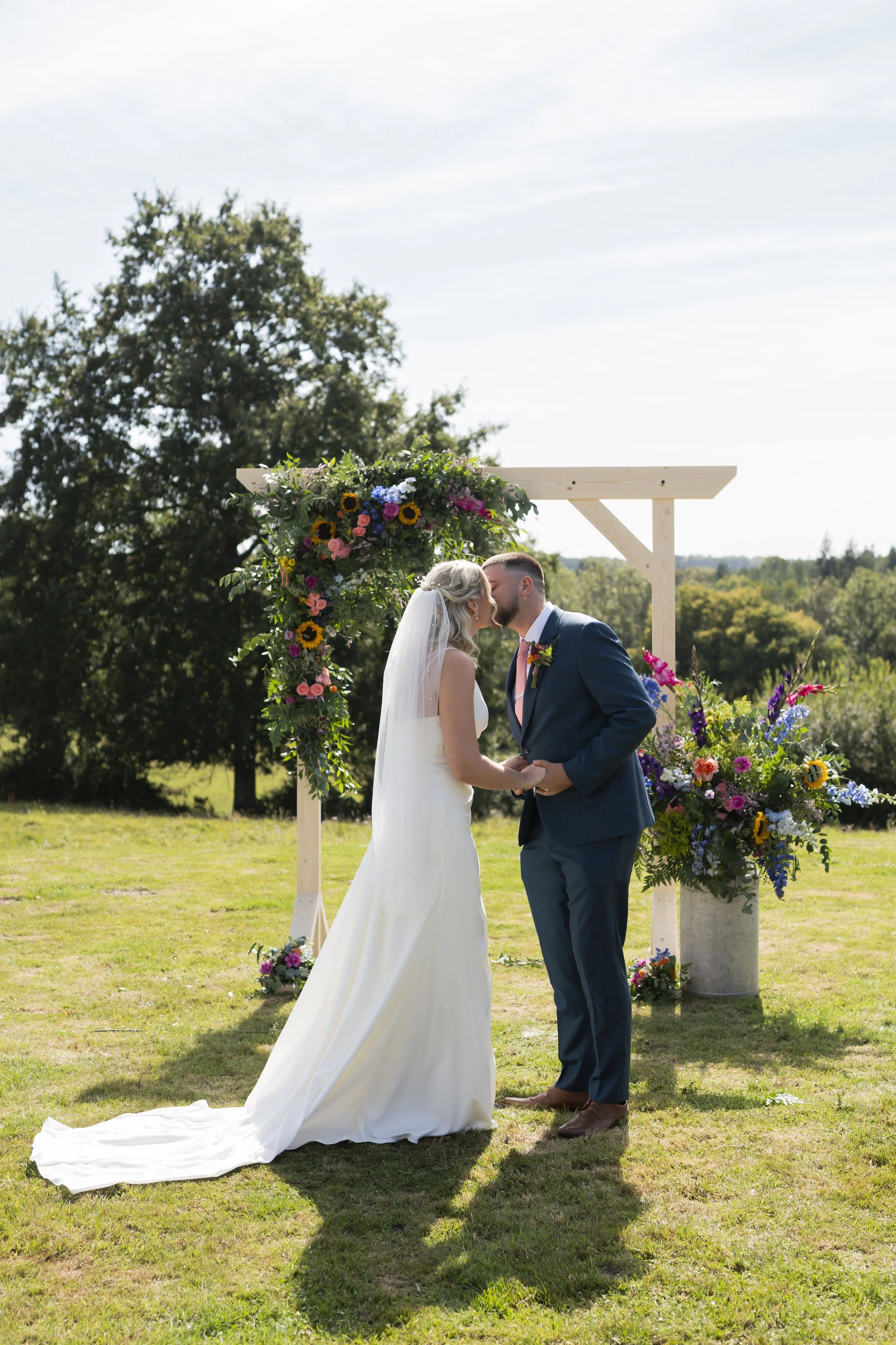 A bride and groom share a kiss under an outdoor wedding arch decorated with colorful flowers, on a sunny day in a grassy field with trees in the background.