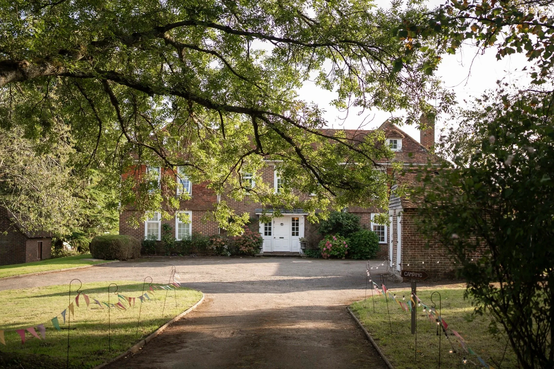 A brick house with a white front door and multiple windows, partially obscured by leafy tree branches overhead. The yard has green grass, bushes, and a gravel driveway with a sign that says 'Camping' and colorful flags on the edges.