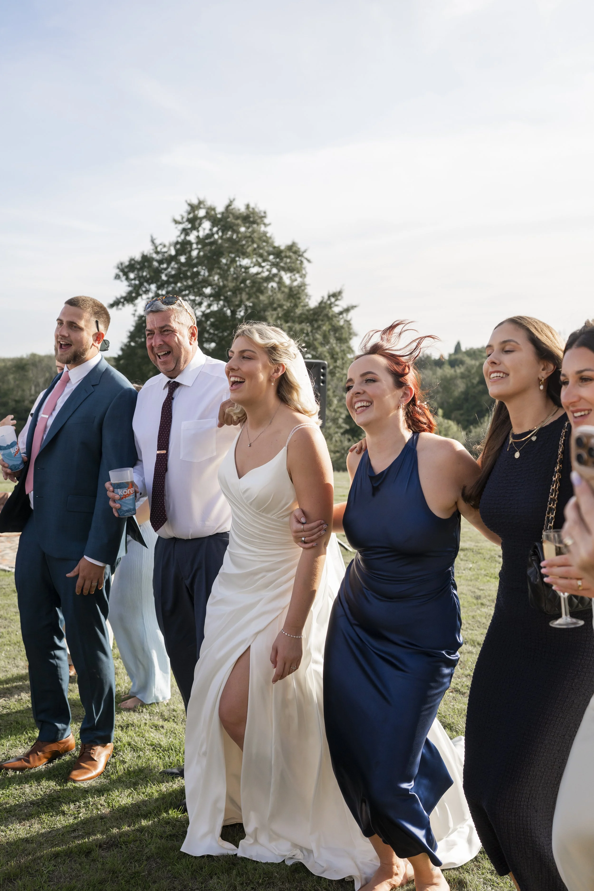 Group of people dressed in formal attire, walking outdoors on grass, smiling and enjoying each other's company, in a sunny setting with trees in the background.