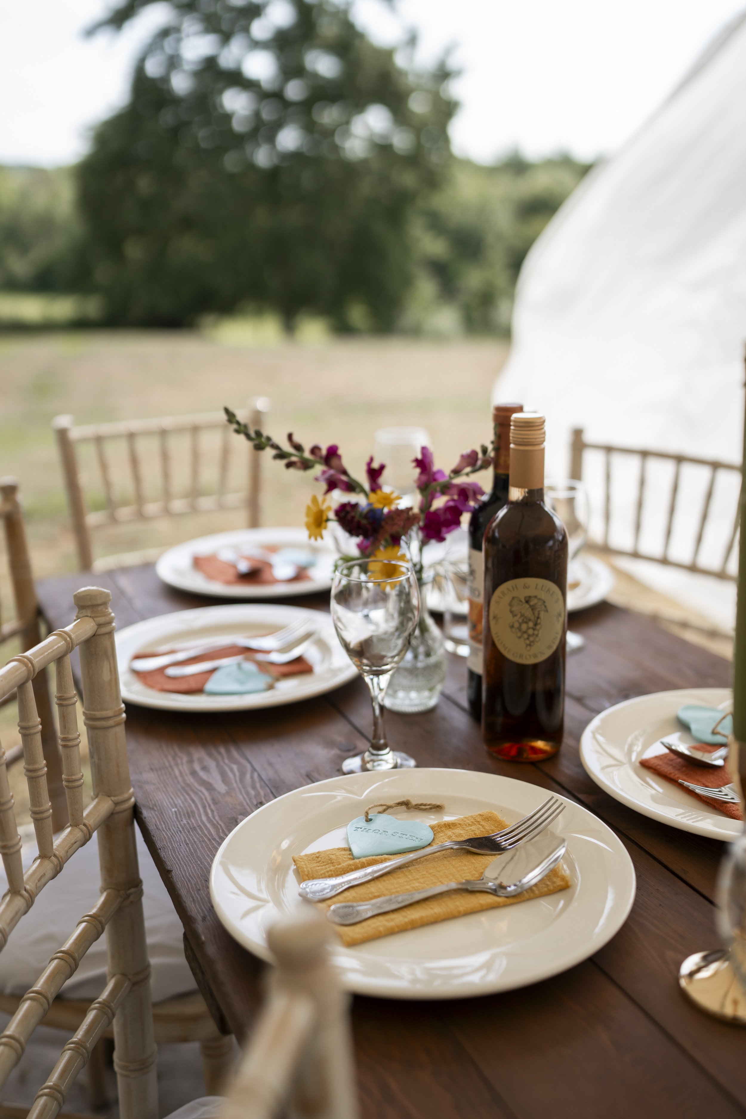 A rustic outdoor dining table set with white plates, cloth napkins, silverware, and wine glasses. Small bottles of wine and a vase with colorful flowers sit in the center, with blurred greenery in the background.