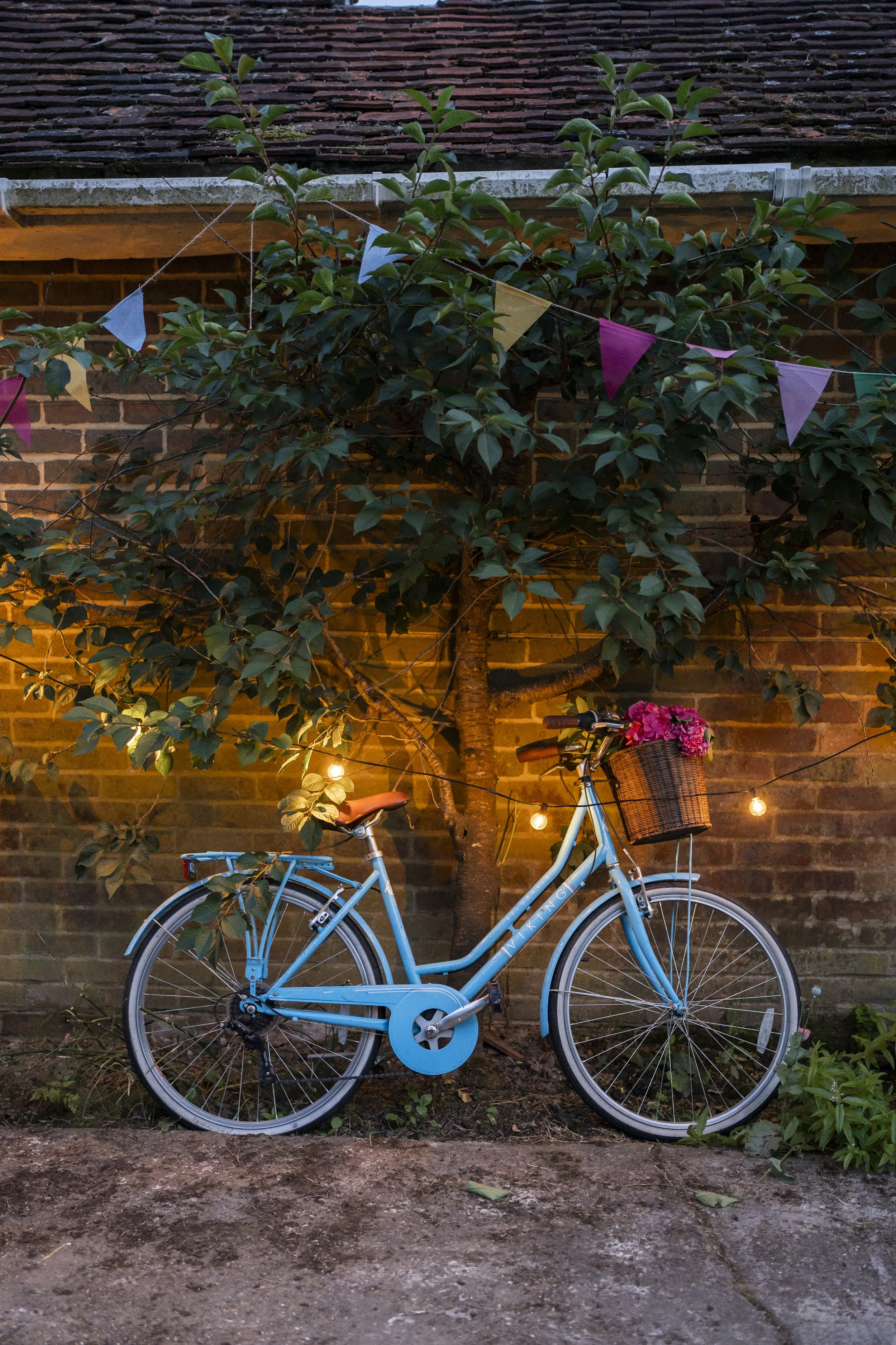 A pastel blue bicycle with a wicker basket filled with pink flowers, parked against a brick wall. There are string lights above, a leafy tree, and a sparse patch of ground in the foreground.