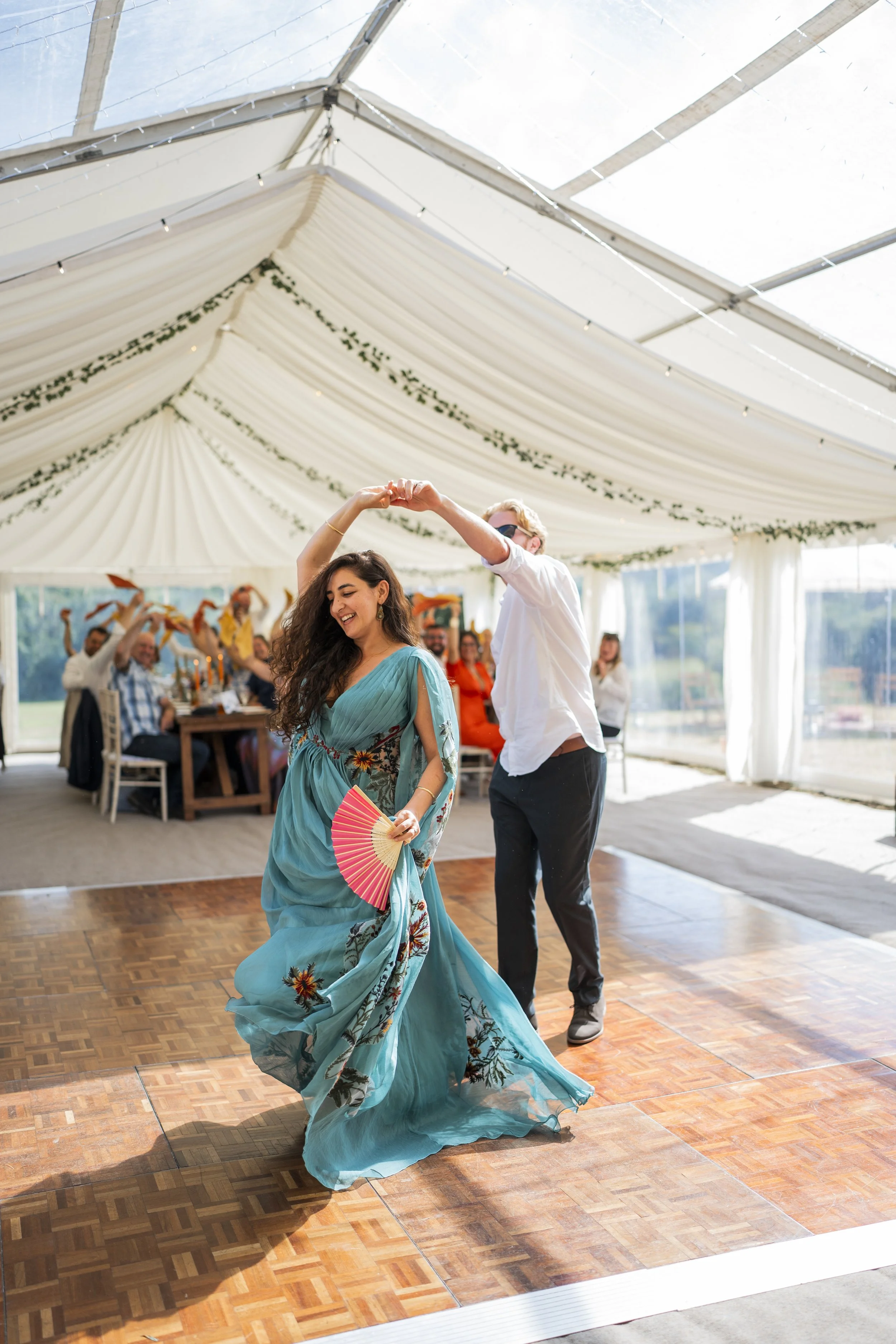 A woman in a blue floral dress dancing at a celebration indoors, with a man in white shirt and sunglasses dancing behind her, and people sitting at tables in the background.