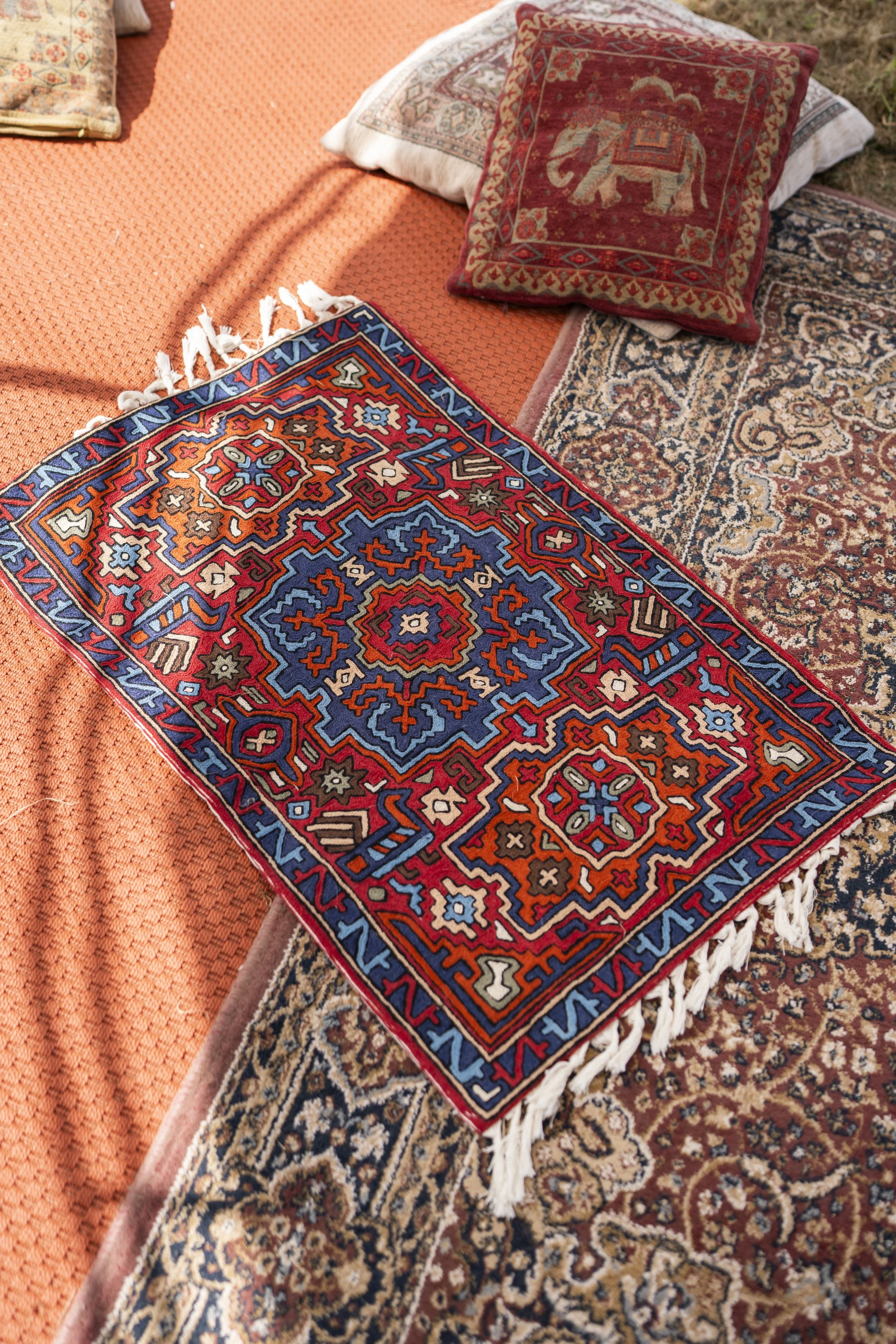 Close-up of colorful, patterned Persian-style rugs and cushions with intricate designs, placed on an outdoor setting with surrounding orange and brown textiles.