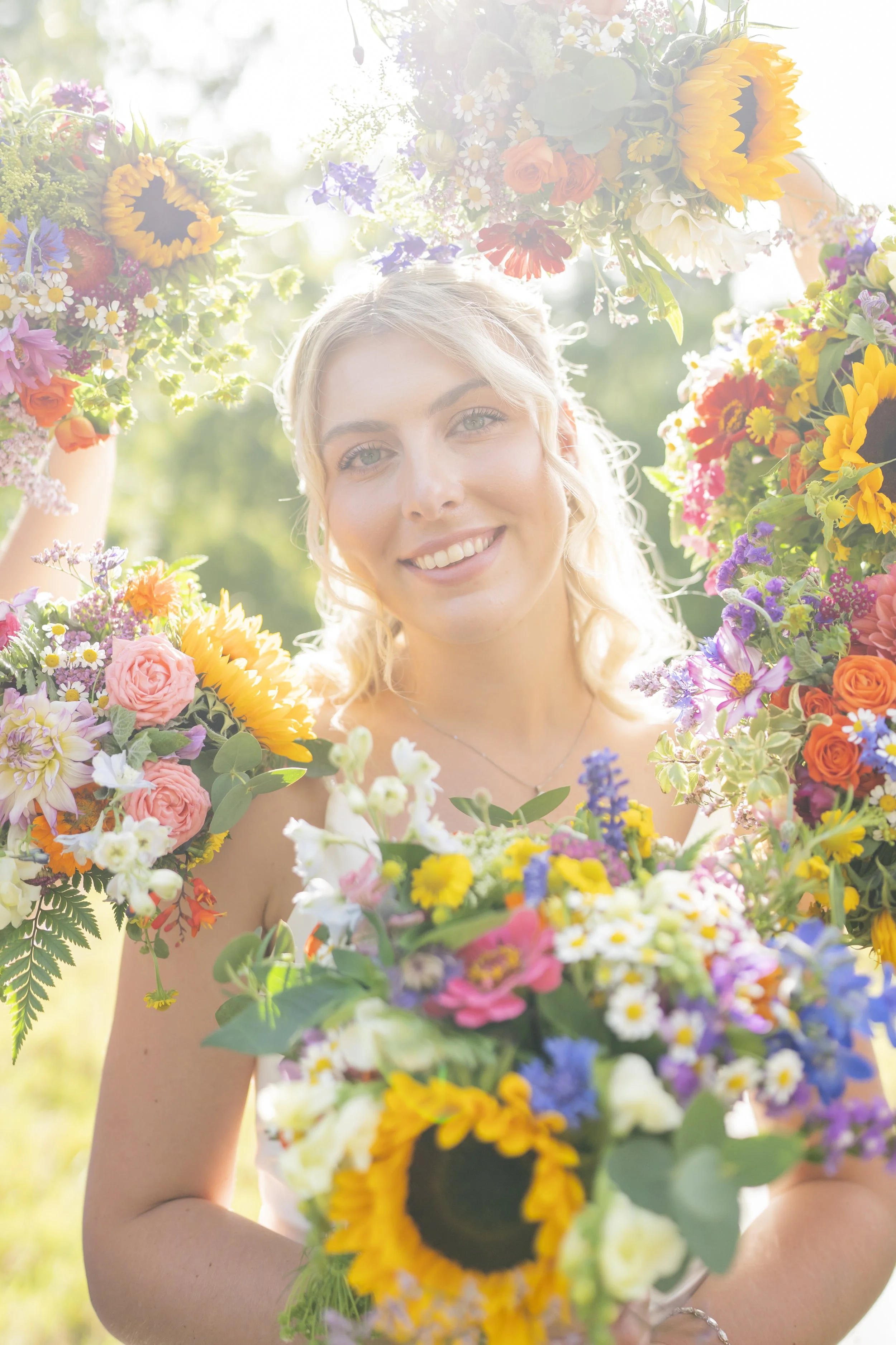 A woman with light hair and blue eyes smiling outdoors amidst a large, colorful assortment of flowers and greenery, with sunlight illuminating the scene.