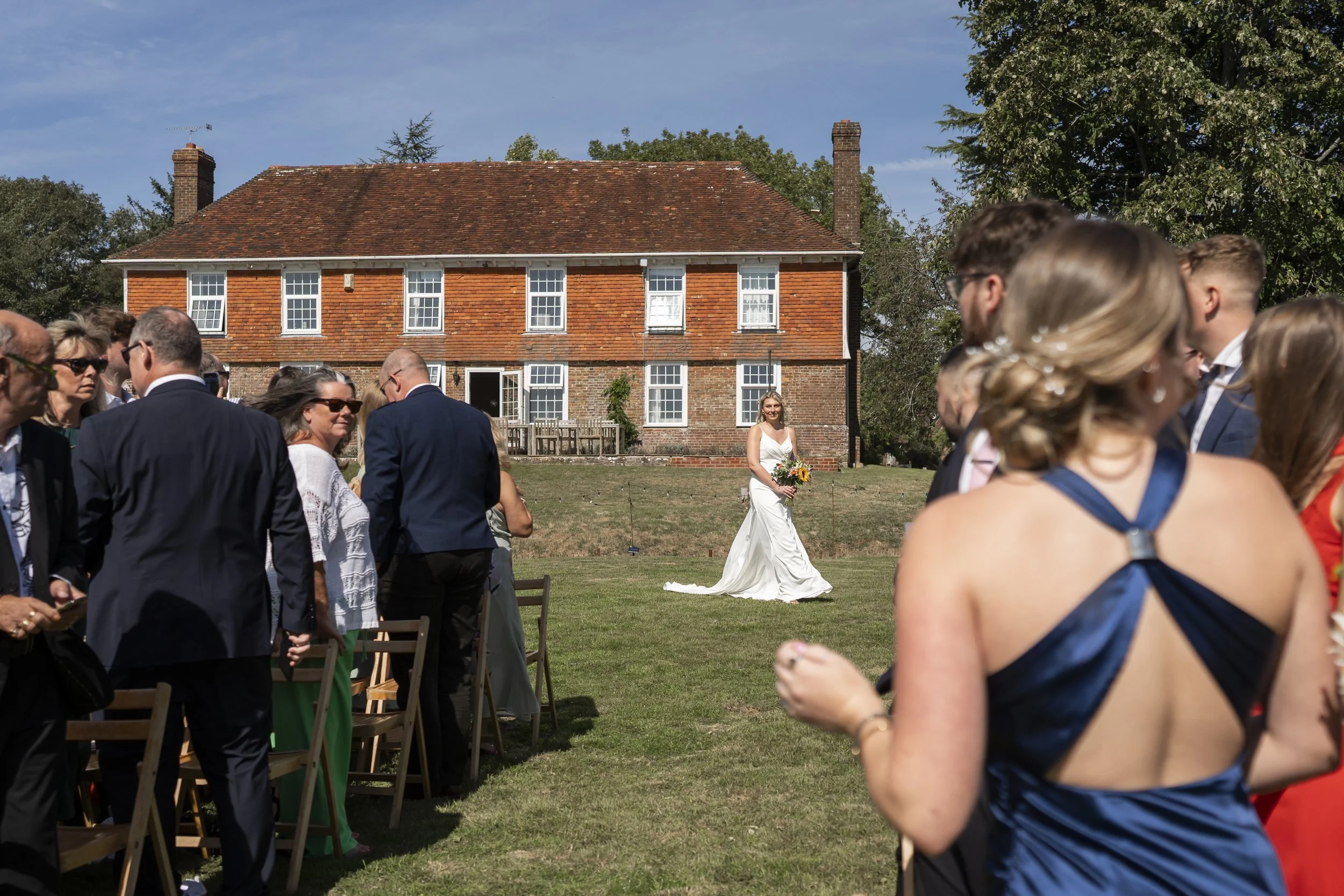A wedding ceremony outdoors on a sunny day with a brick house in the background. A bride in a white gown and holding a bouquet stands in the middle, surrounded by guests dressed in formal attire.