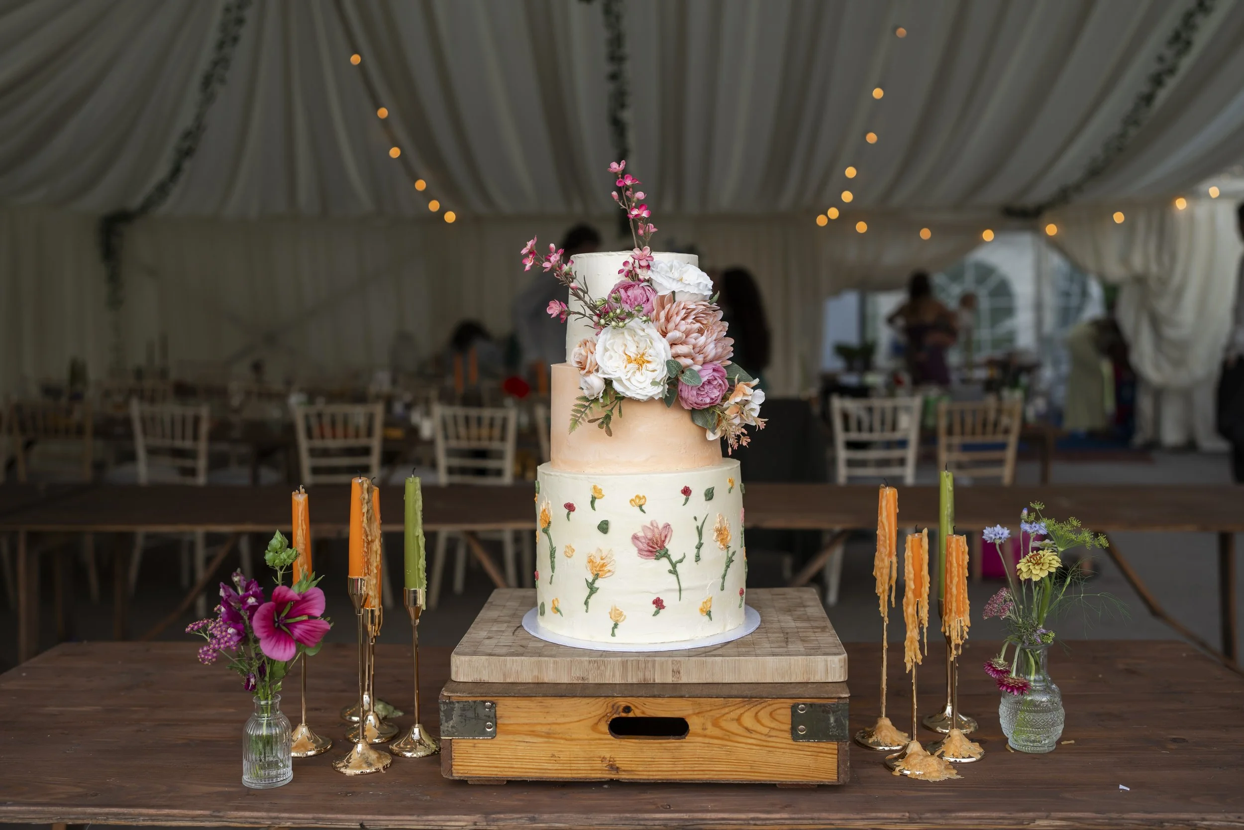 Decorative wedding cake with floral decorations on a wooden table, surrounded by colorful candles and flower arrangements, in a decorated venue.