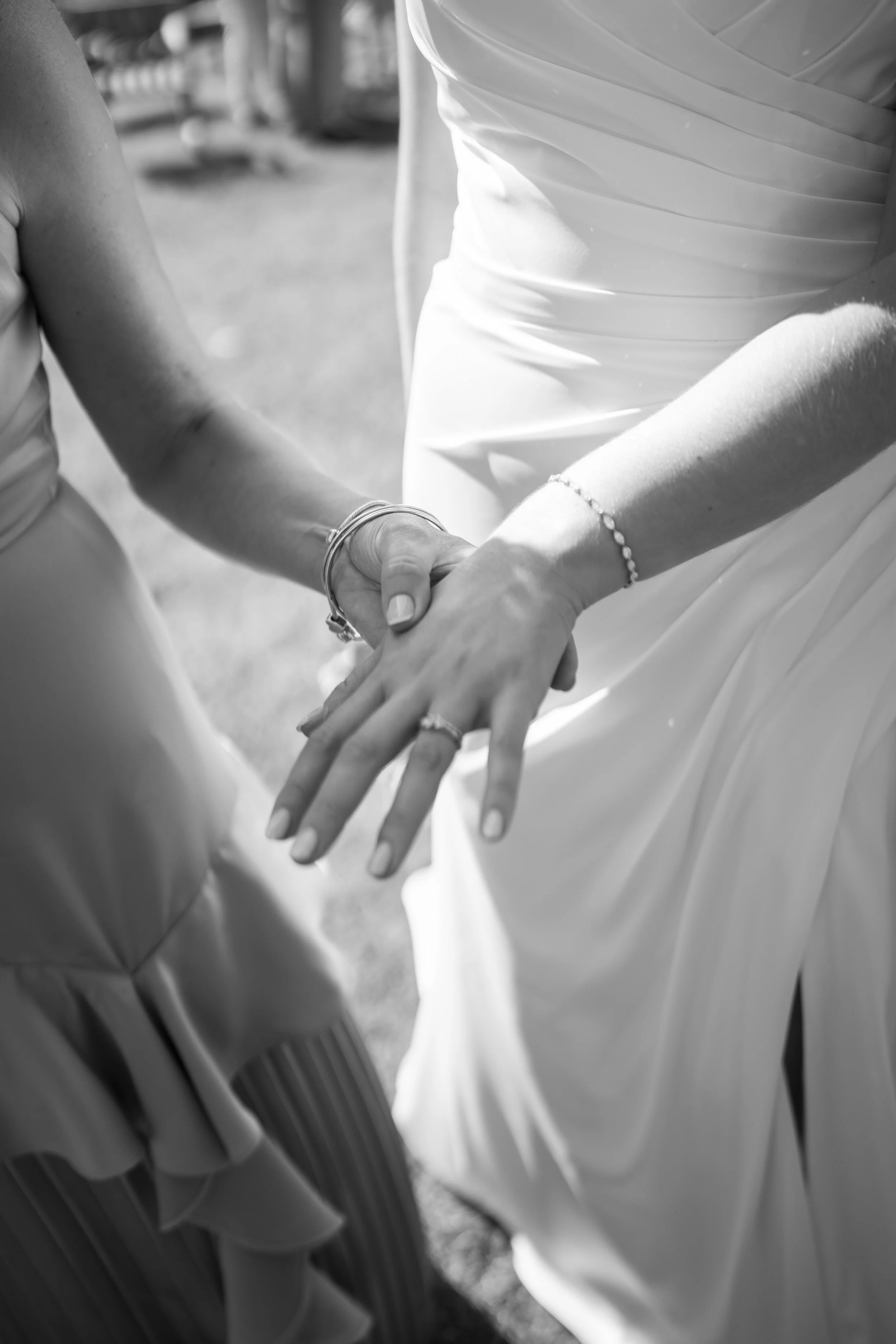 Two women in wedding dresses holding hands, dressed in white, with bracelets on their wrists, outdoors.