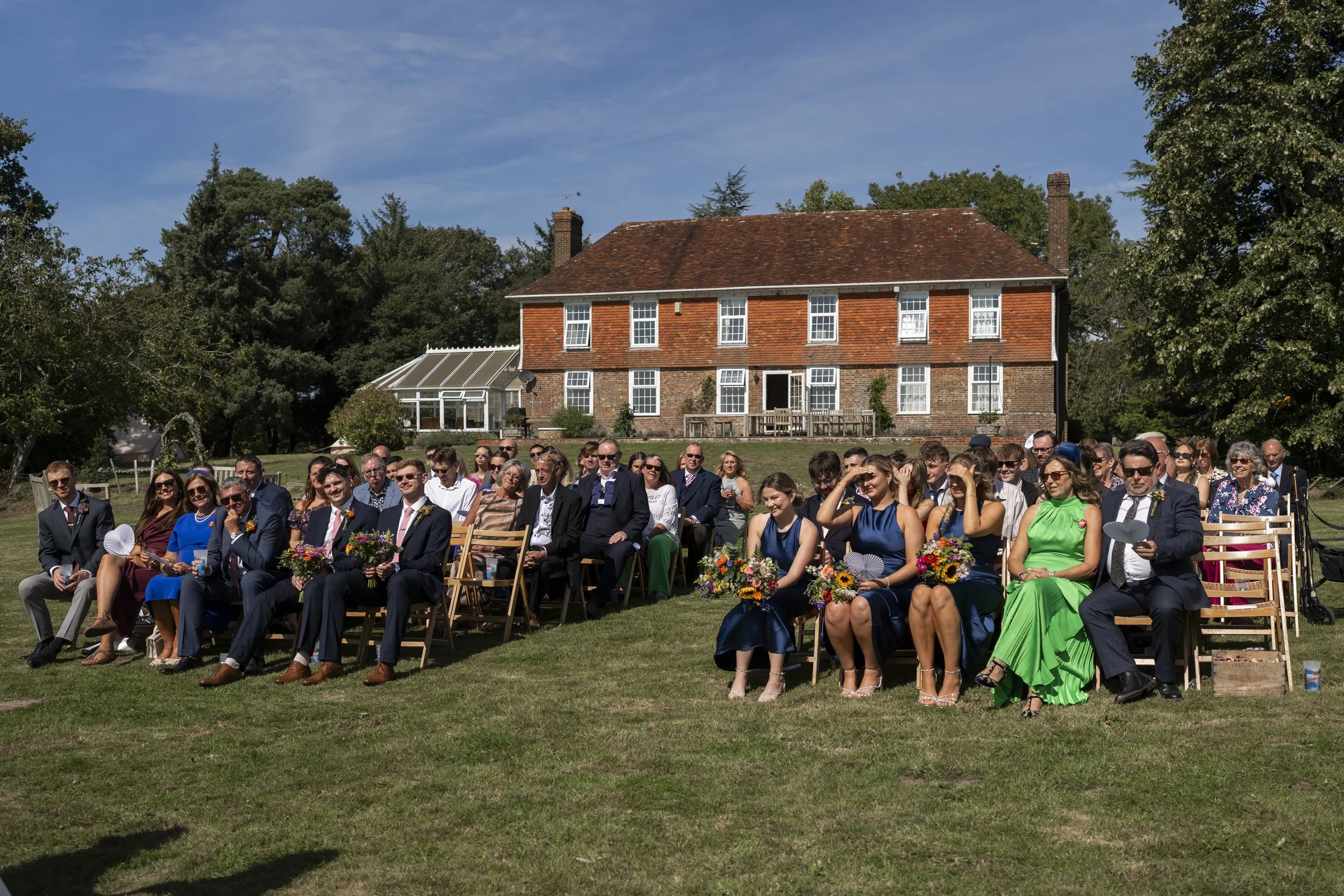 A large group of people attending a wedding or outdoor event seated on chairs on a grassy lawn with a large brick house and trees in the background. The people are dressed in formal attire and appear to be enjoying the occasion.